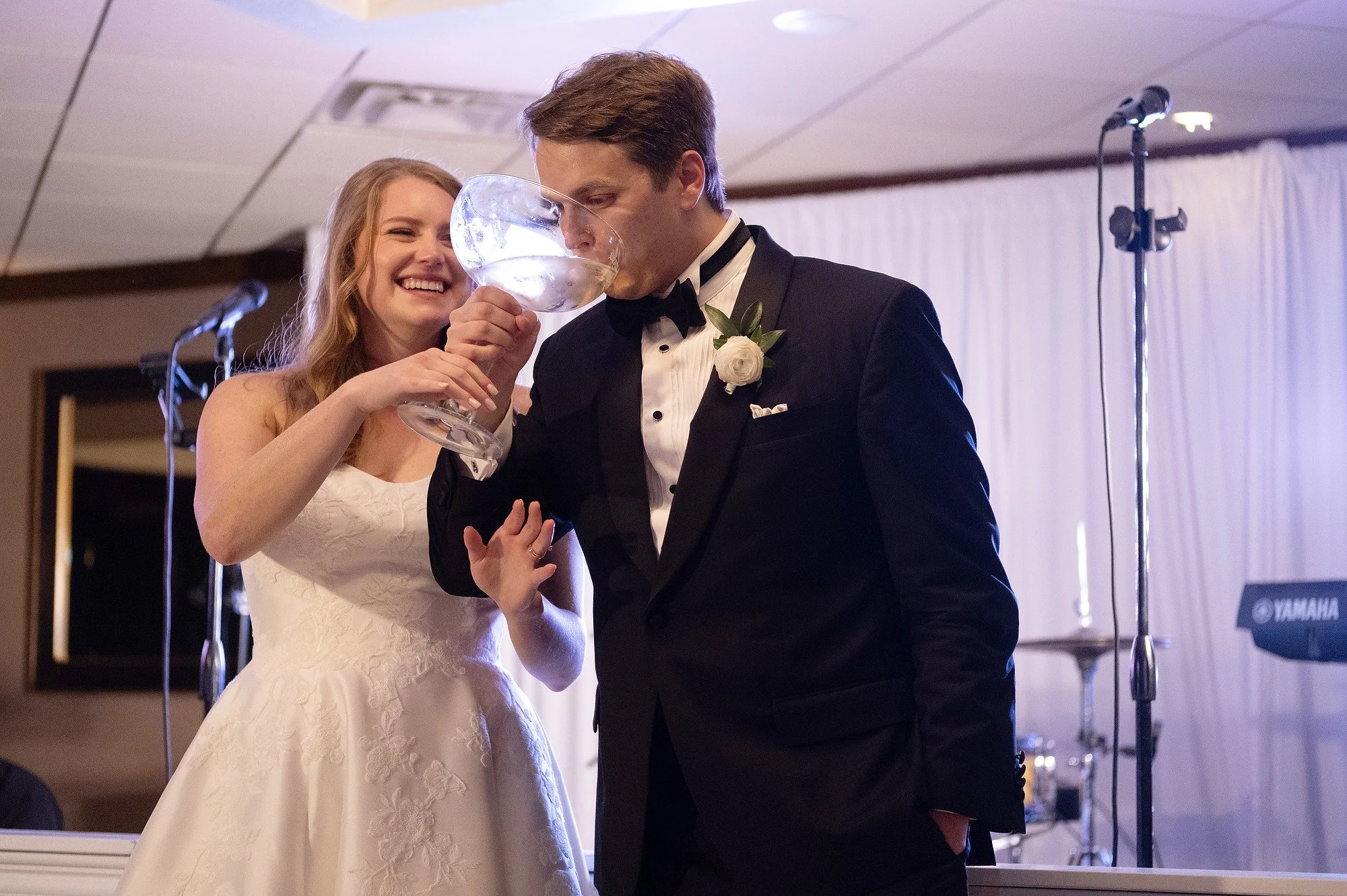 A bride and groom at their wedding reception, with the bride holding a large glass, about to take a sip, while the groom stands close with his eyes closed. The bride is smiling and wearing a white wedding dress, and the groom is dressed in a black tu