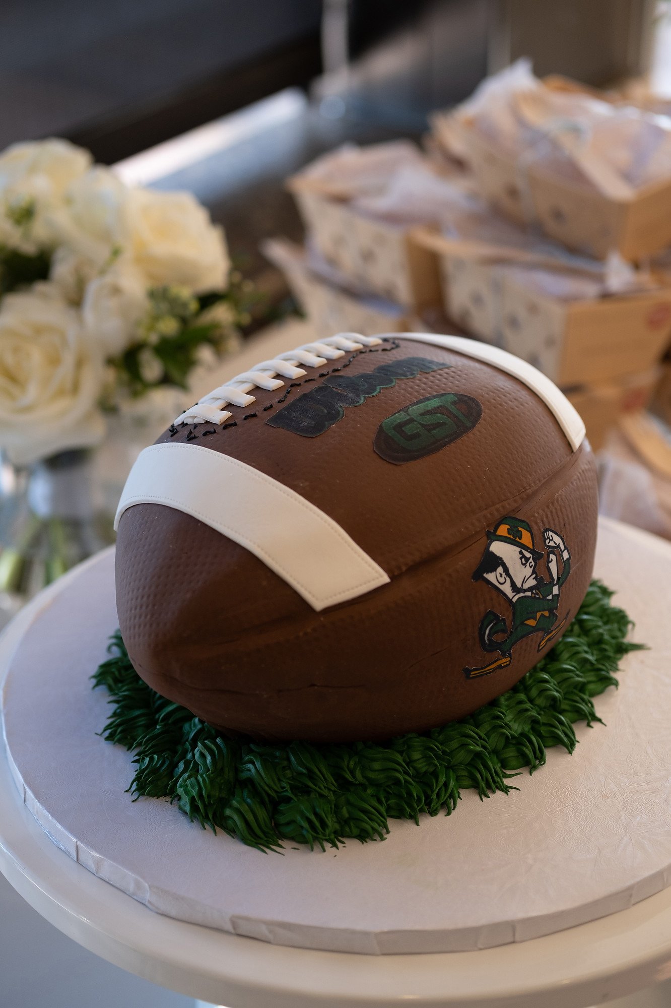 Football-shaped cake decorated with football markings and team logos, placed on a white cake stand with green grass-like icing around its base.