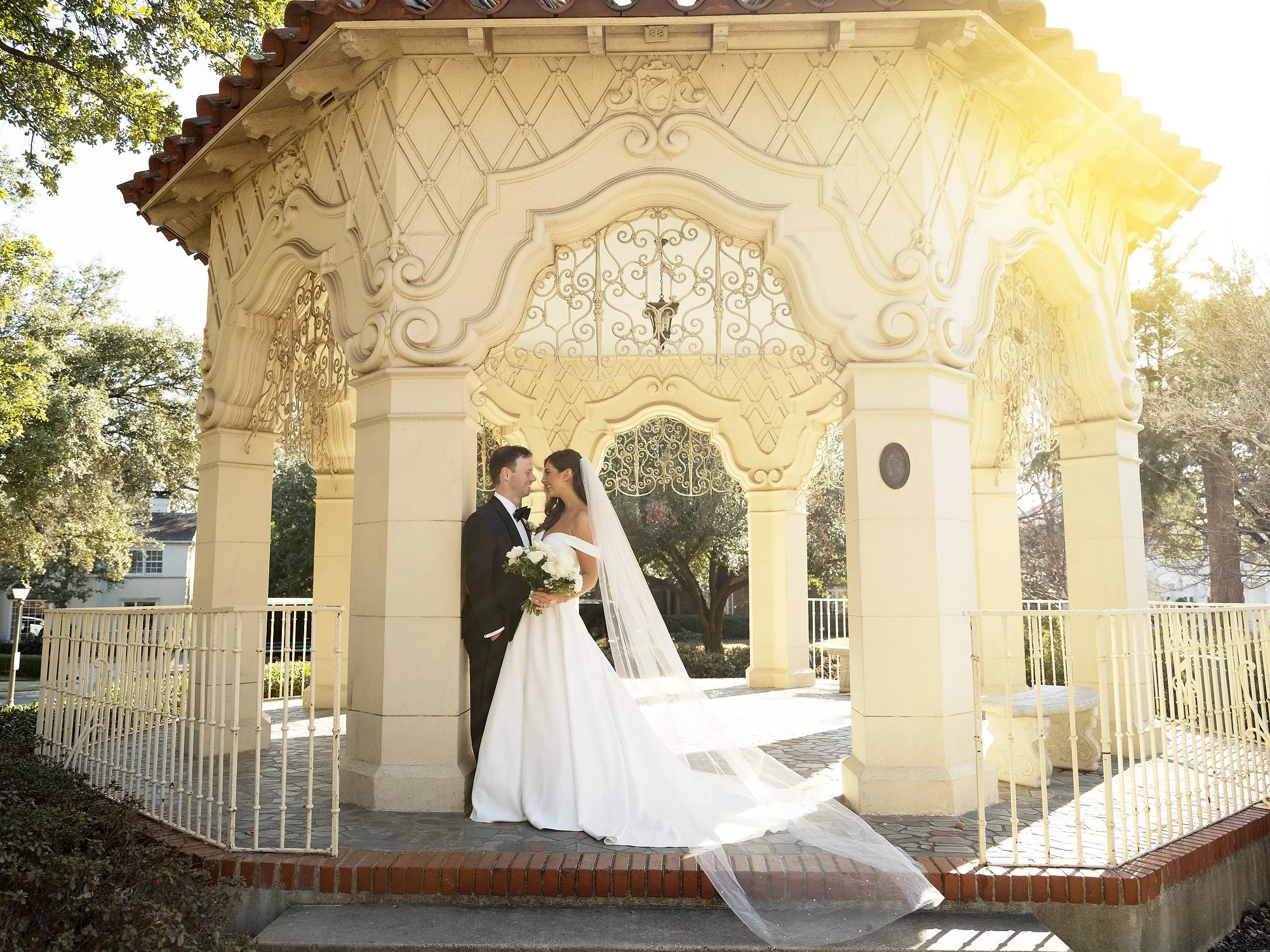 A bride and groom in wedding attire standing close together under a decorative gazebo, with the bride holding a bouquet of white flowers, outdoors during daytime with sunlight filtering through trees.