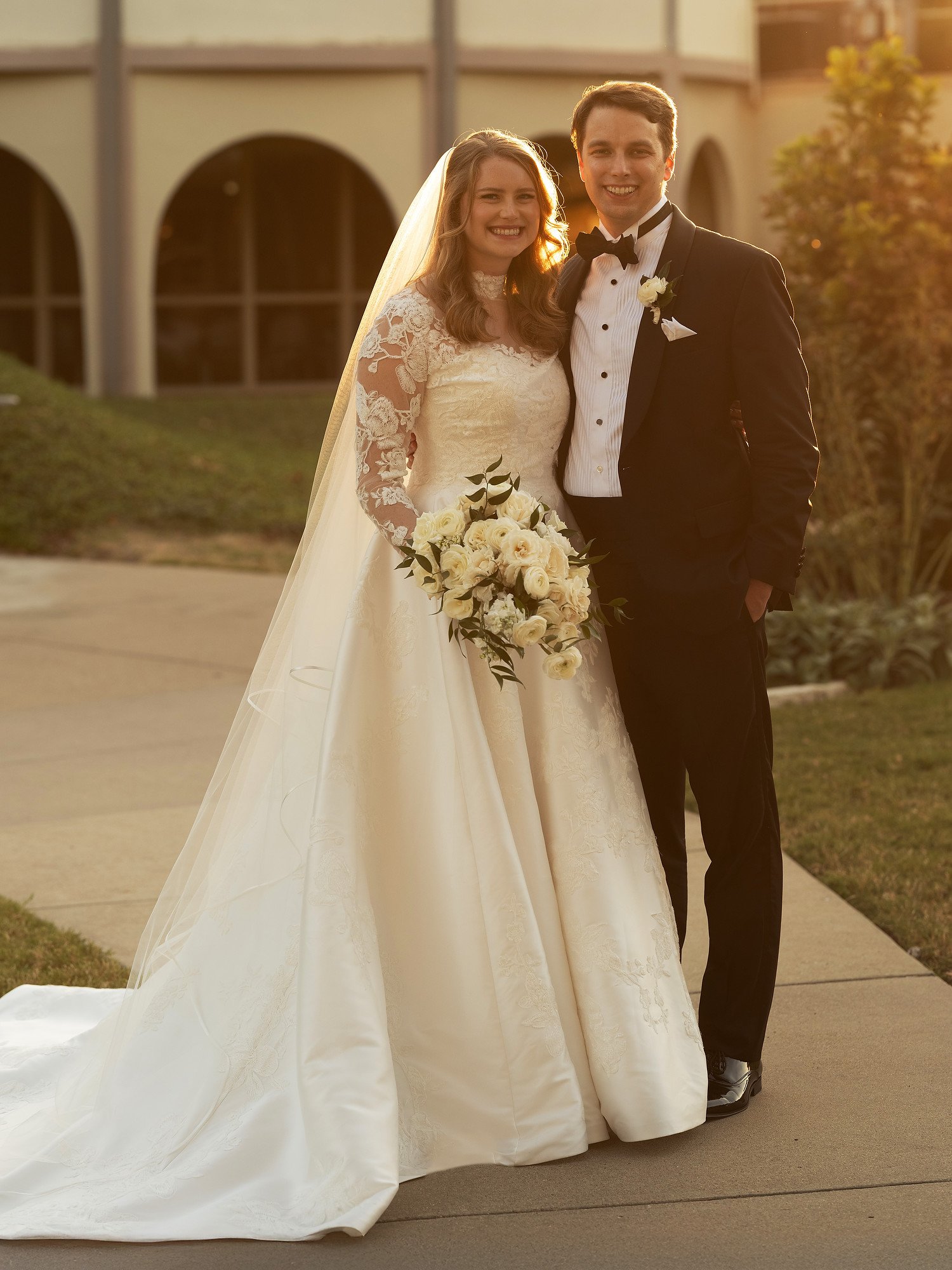 A bride and groom posing outdoors on their wedding day at sunset, both smiling. The bride is wearing a lace wedding gown with a veil and holding a bouquet of white roses. The groom is dressed in a black tuxedo with a bow tie and a white boutonniere. 