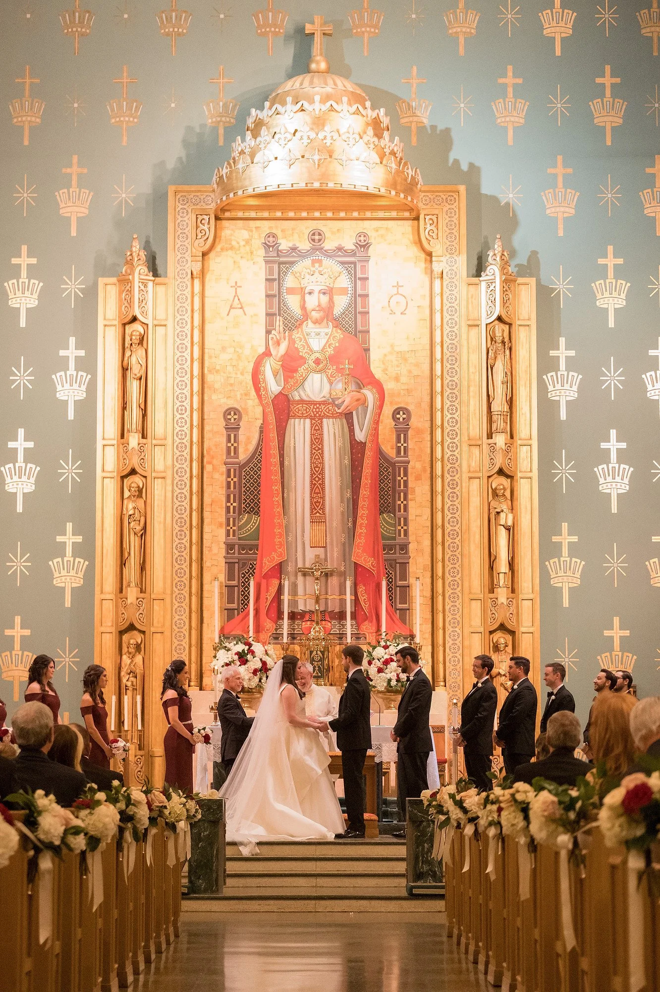 A wedding ceremony taking place inside a church with a large religious icon of Jesus Christ in the background, surrounded by ornate woodwork and statues. The bride and groom are standing at the altar, exchanging vows, with bridesmaids and groomsmen o