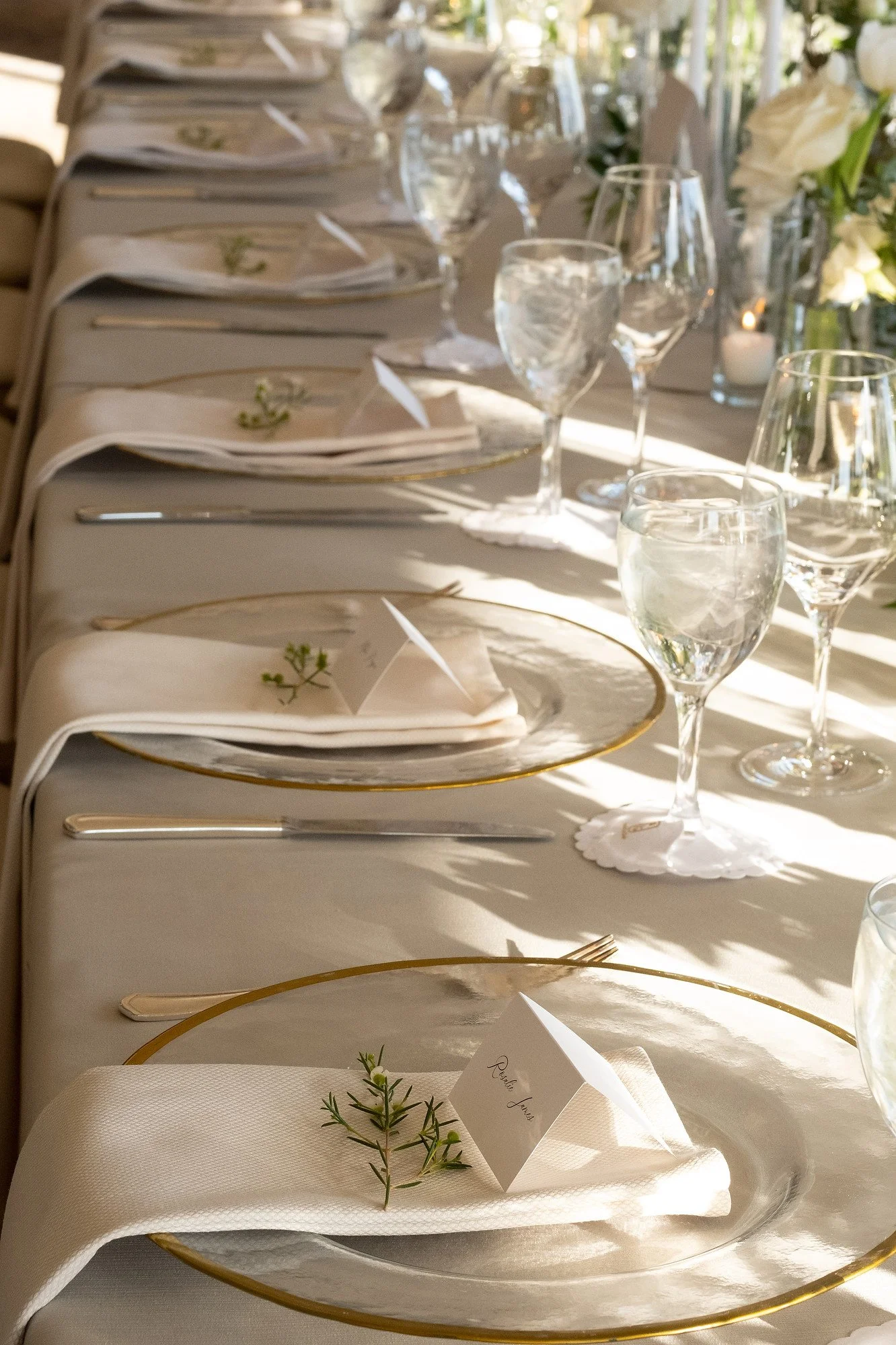 Elegant banquet table set for a formal event with white tablecloths, clear glassware, white napkins decorated with small green sprigs, and folded name cards in front of each seat, illuminated by natural light and surrounded by white floral arrangemen