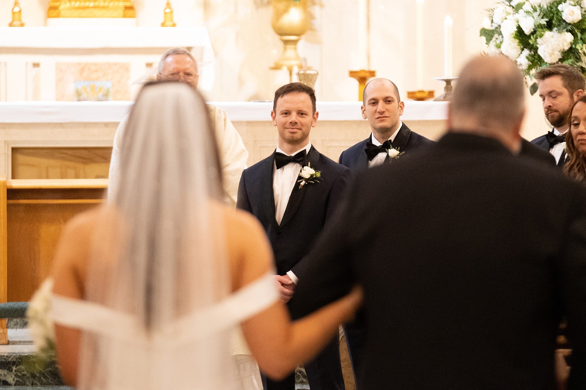 A bride and groom stand at the altar during their wedding ceremony, facing a priest, with the bride's back to the camera and the groom smiling in a tuxedo. Other wedding guests are visible in the background.
