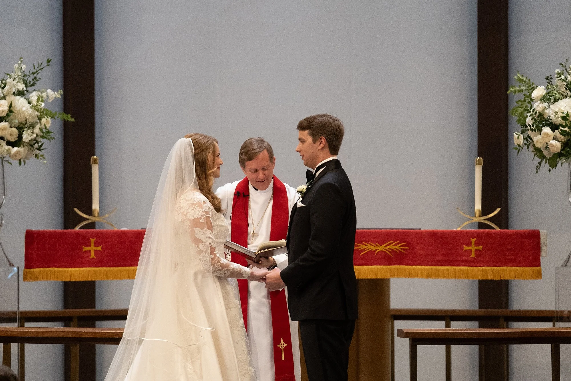 A bride and groom exchanging vows with a minister during a church wedding ceremony, holding hands and standing in front of an altar decorated with candles and flowers.