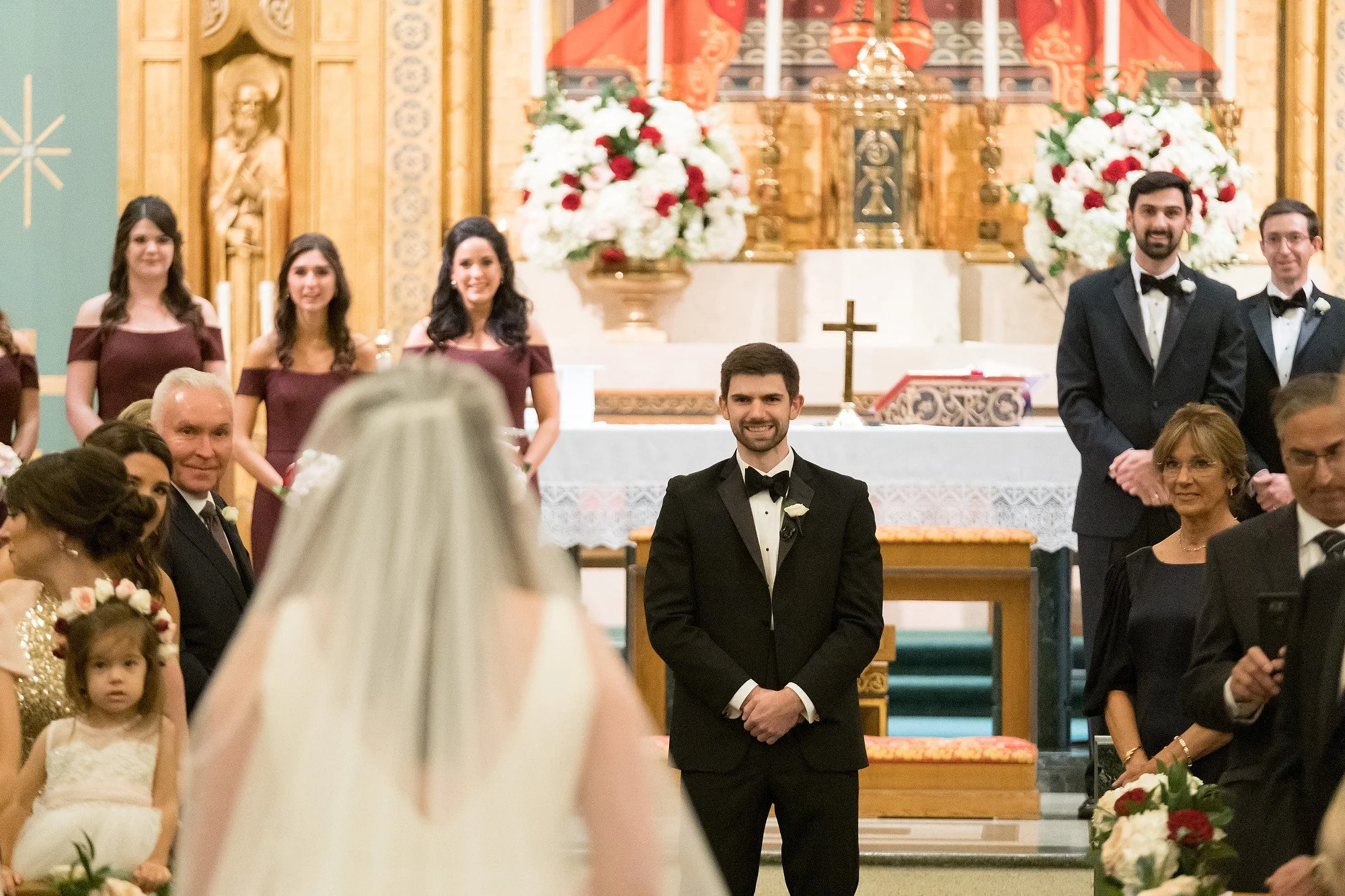 Groom smiling during his wedding ceremony inside a church, with family and friends seated around, an altar with flowers and religious artifacts in the background.