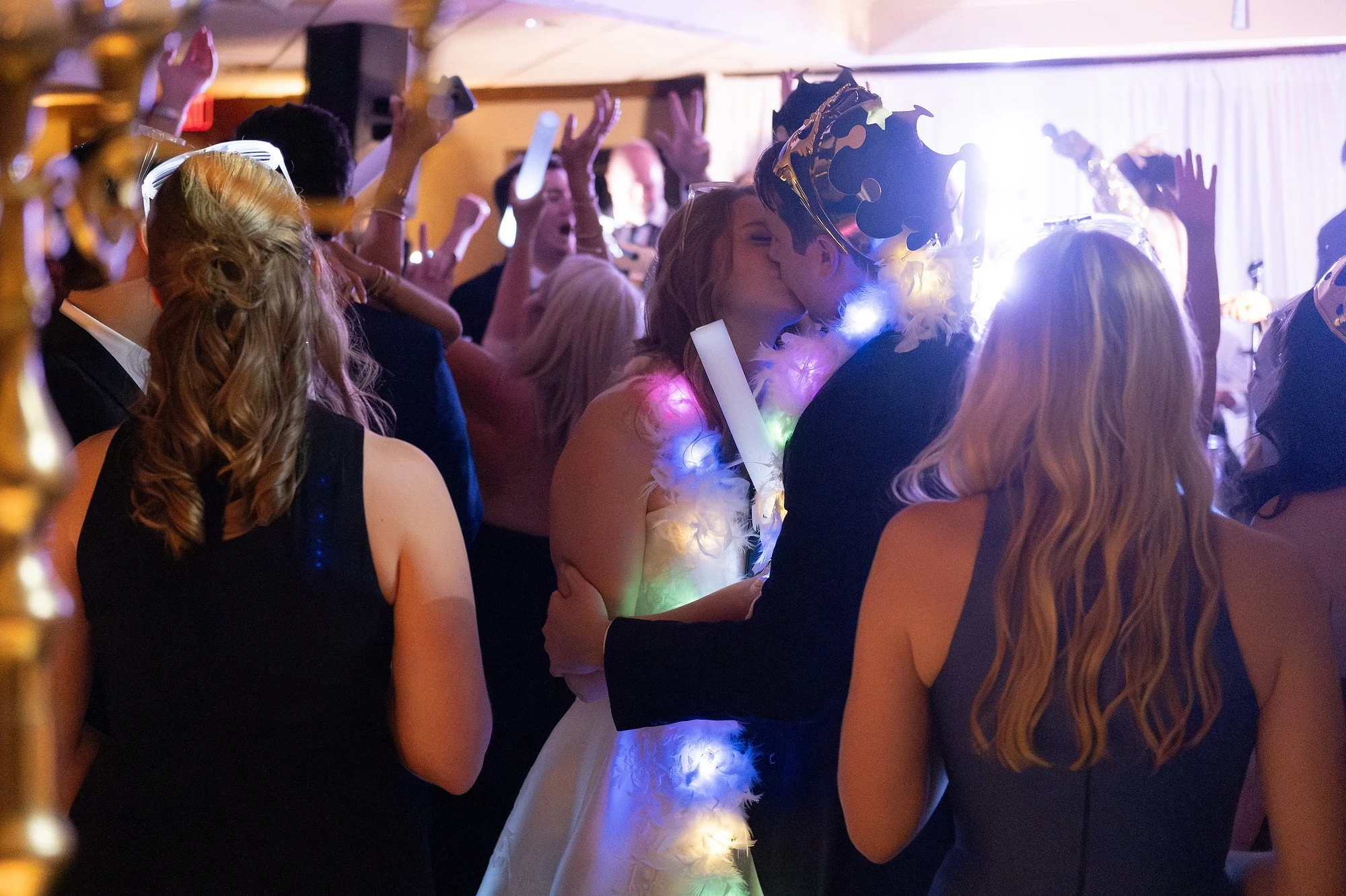 Couple wearing a crown and feather boa kissing at a crowded celebration, with people dancing and raising their hands in the background.