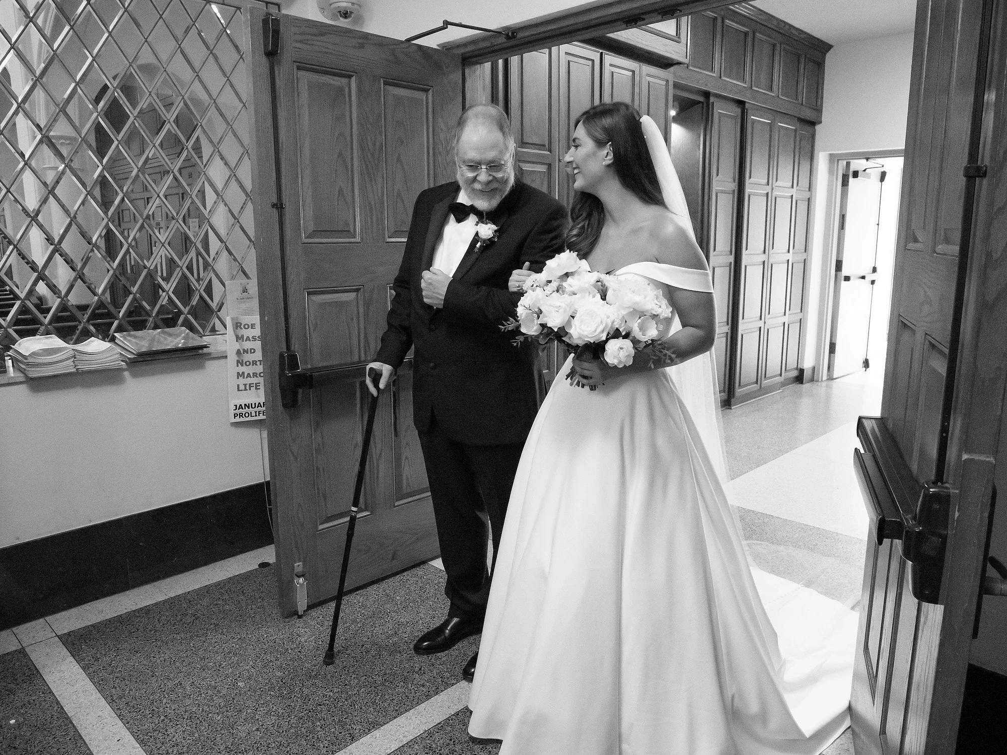 A bride in a wedding dress holding a bouquet of flowers walking with a man in a tuxedo with a cane in a hallway.