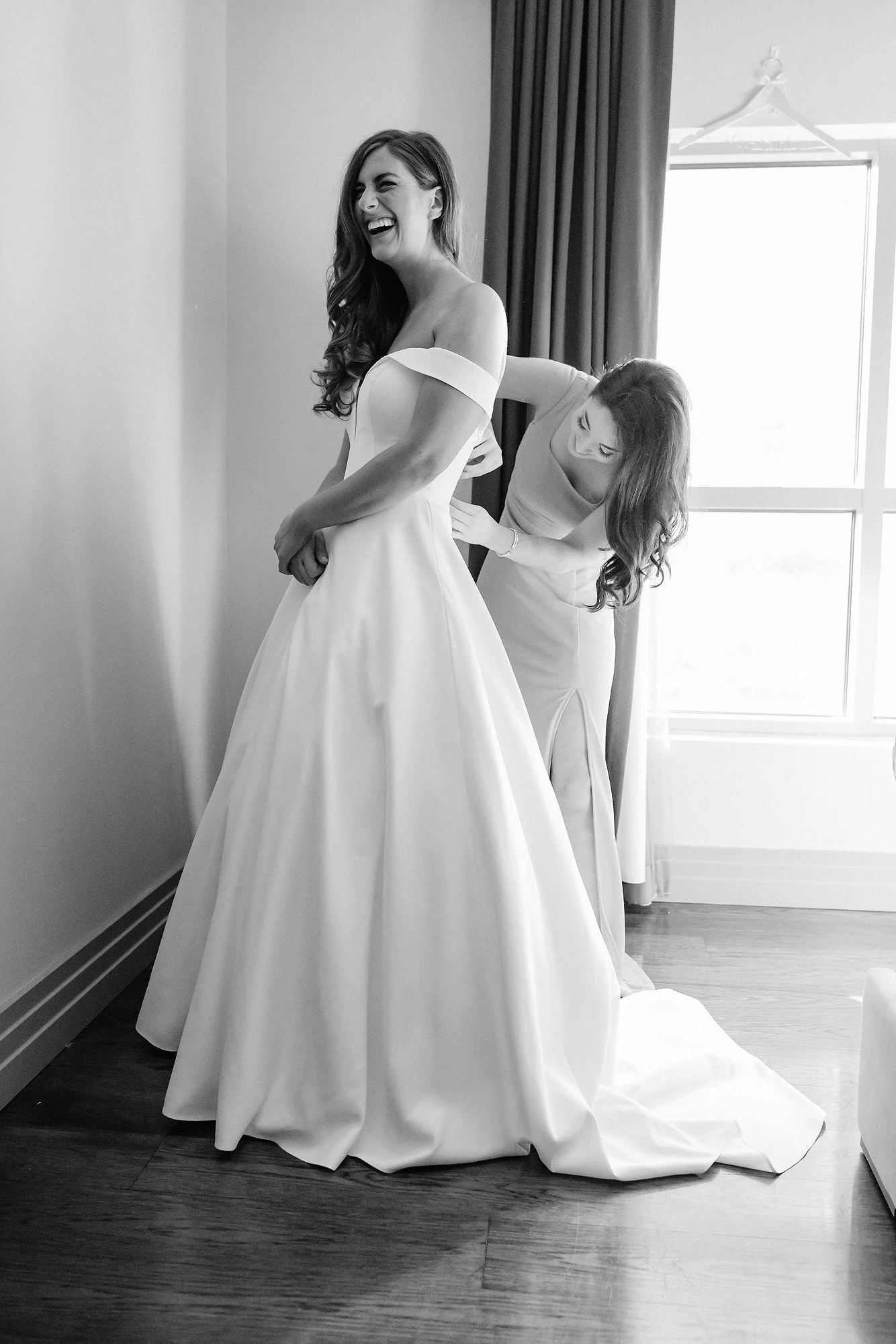 A bride in a wedding dress being assisted by a woman, in a room with sunlight coming through a window, preparing for her wedding.