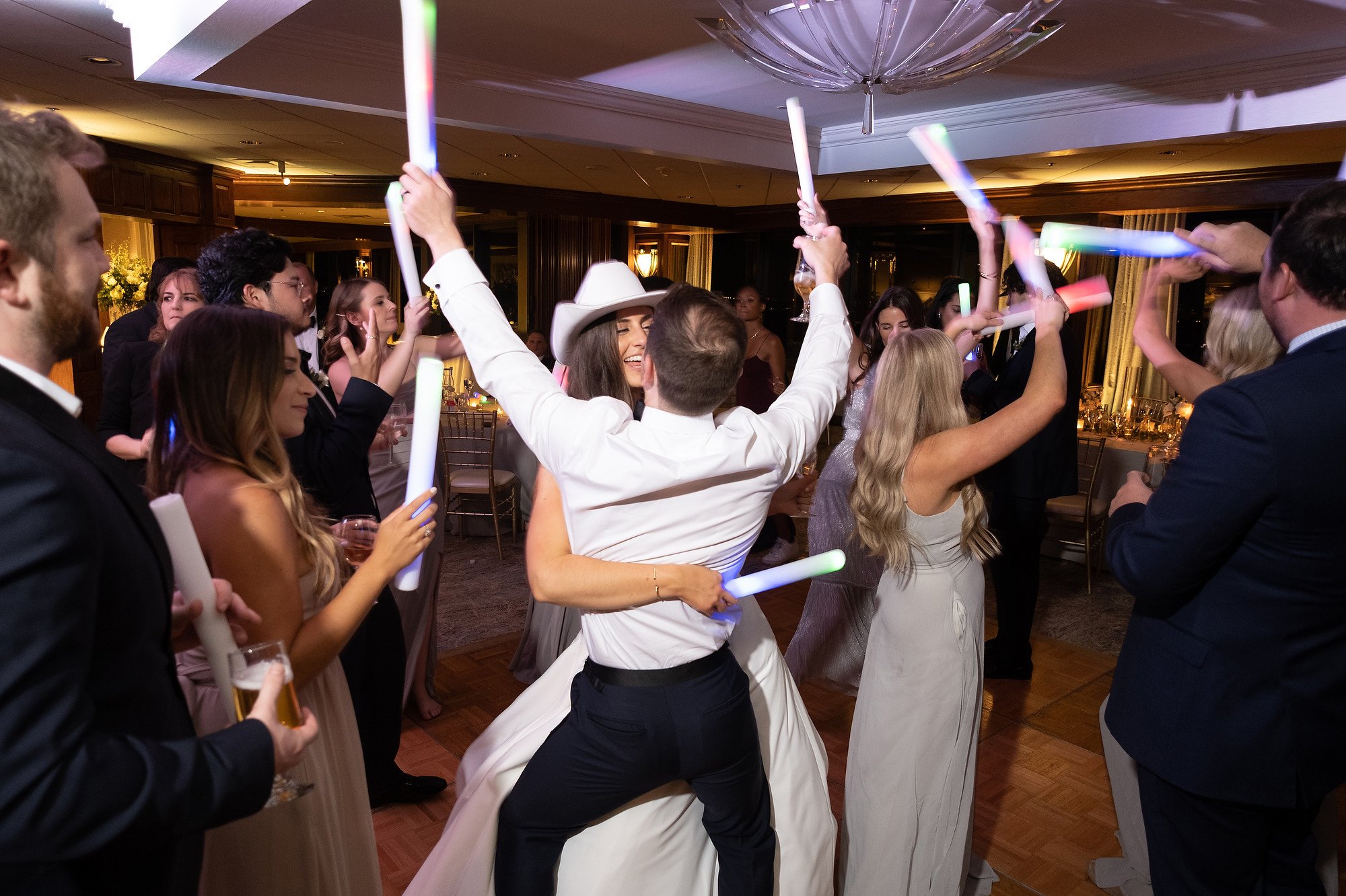A group of people celebrating at a wedding reception, dancing with glow sticks and raising their hands in celebration.