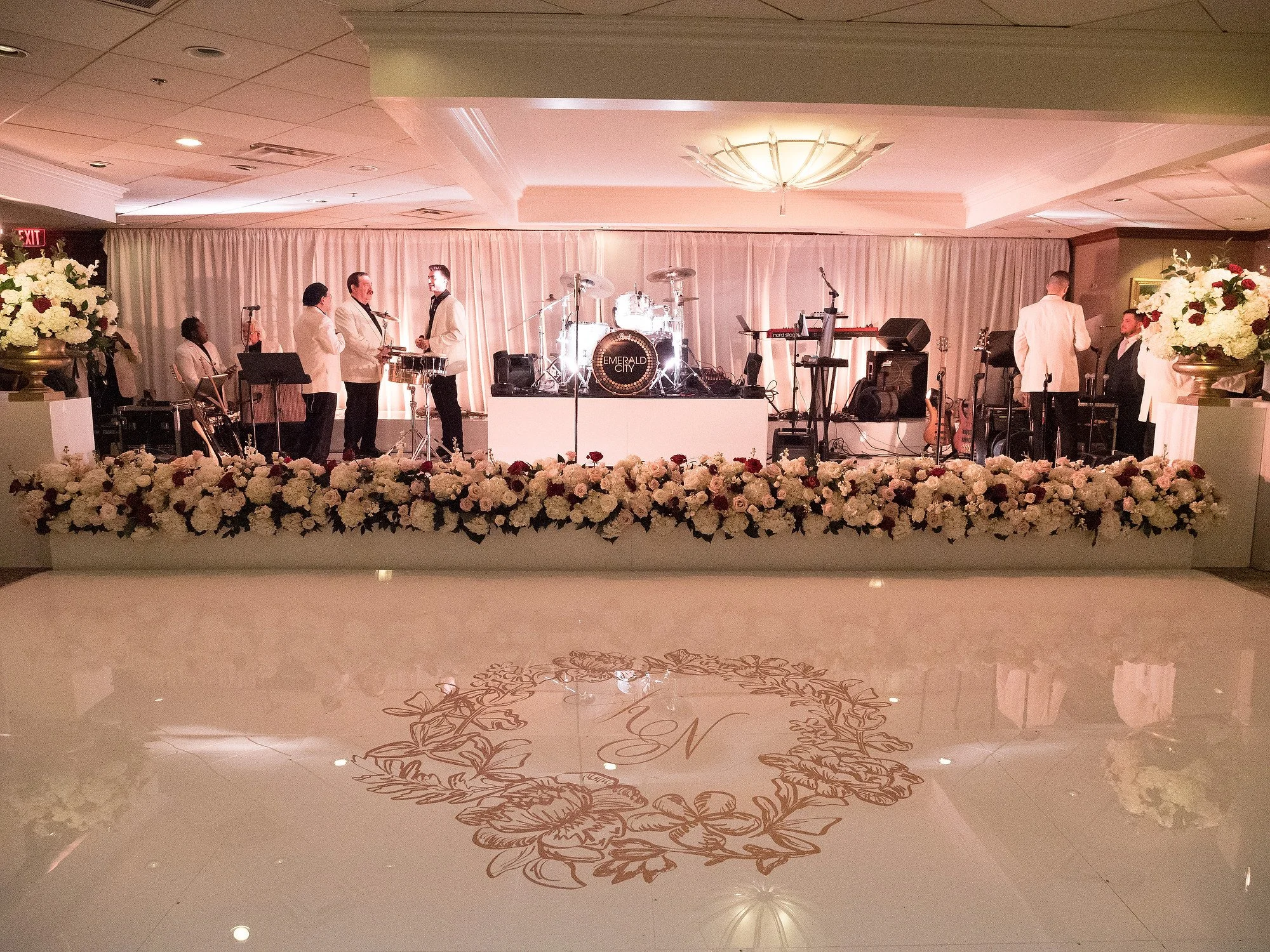 A wedding reception stage decorated with white and pink flowers, with a musical band in white tuxedos setting up instruments and microphones.