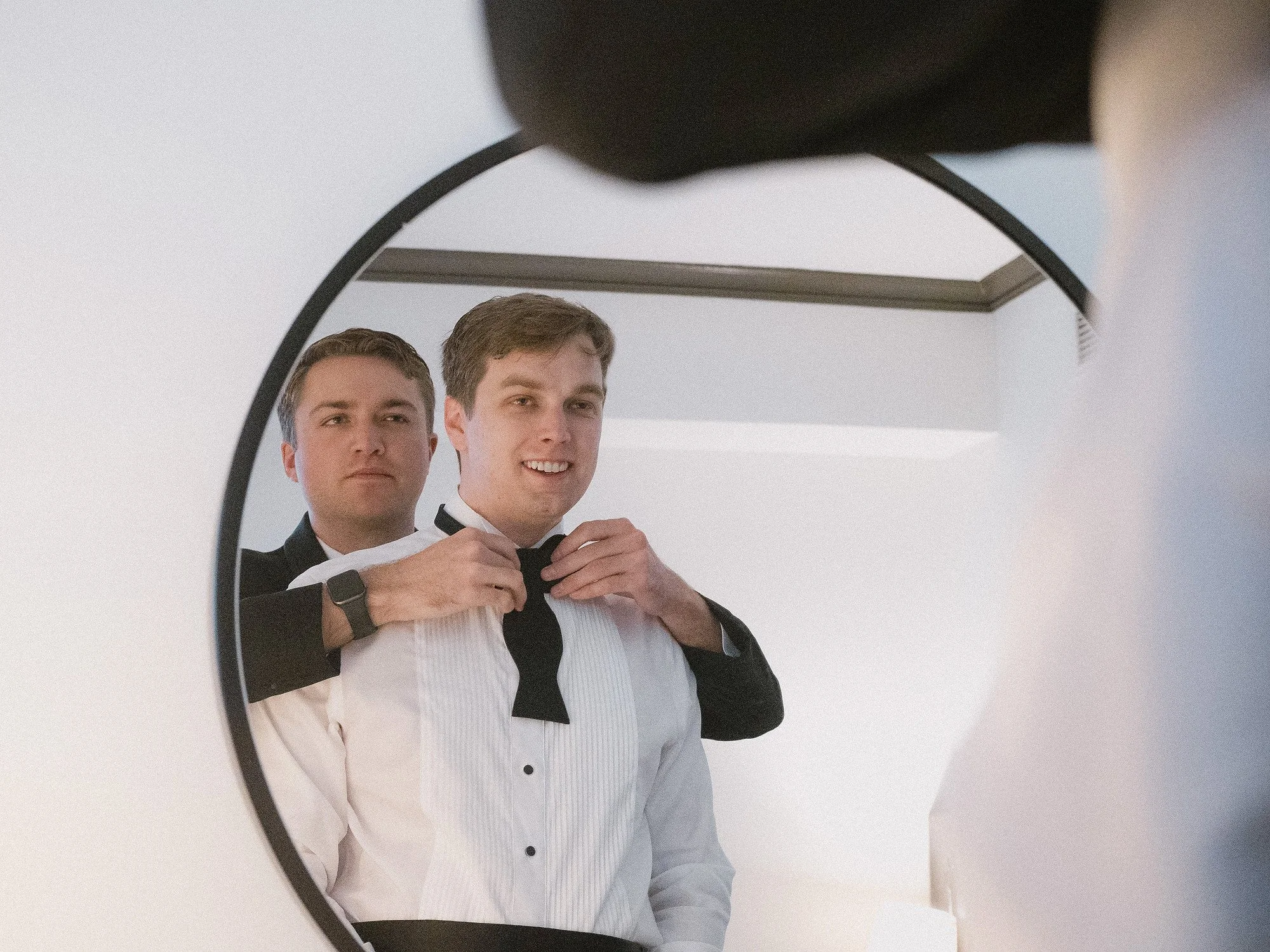 Two men in formal suits, one helping the other adjust his bow tie, as seen in a mirror.