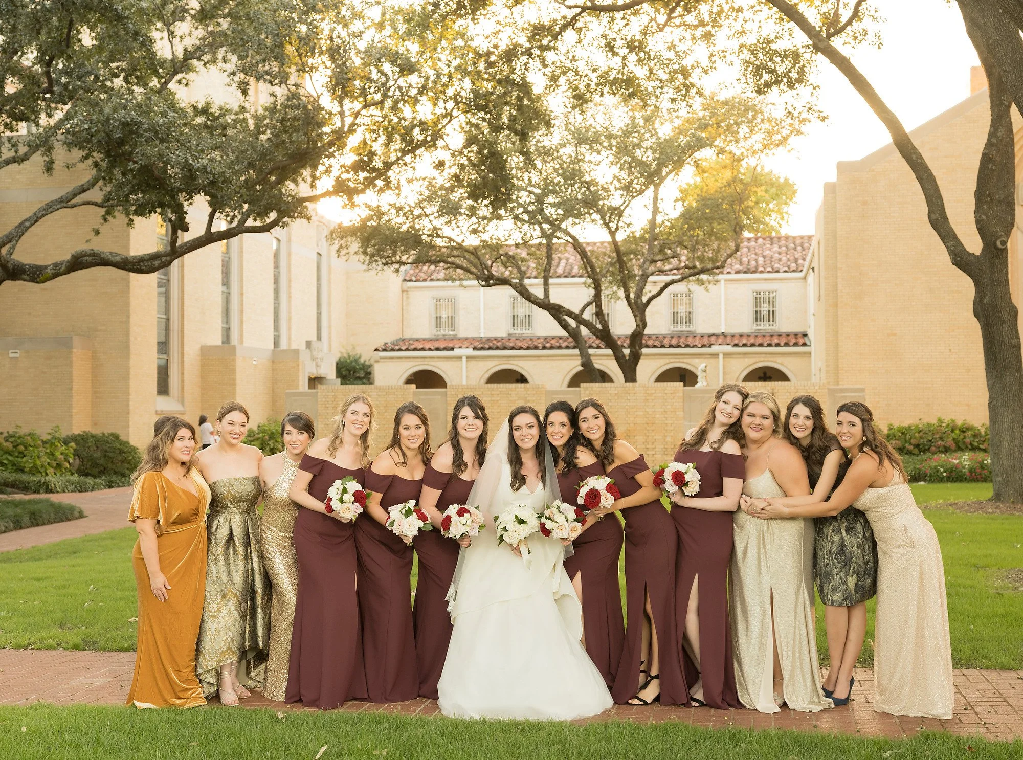 A group of women, including a bride in a white wedding gown holding a bouquet, posing outdoors with bridesmaids in burgundy dresses holding bouquets, standing on a brick pathway with grass and trees in the background during sunset.