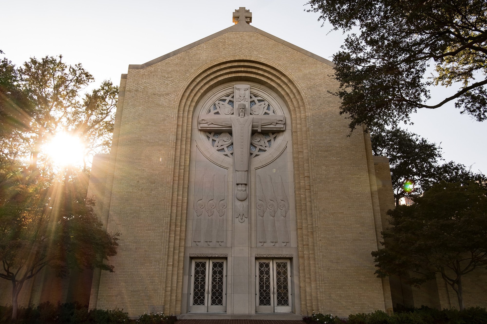 Church with a large bas-relief sculpture of Jesus Christ on the front facade, with sunlight shining through surrounding trees.