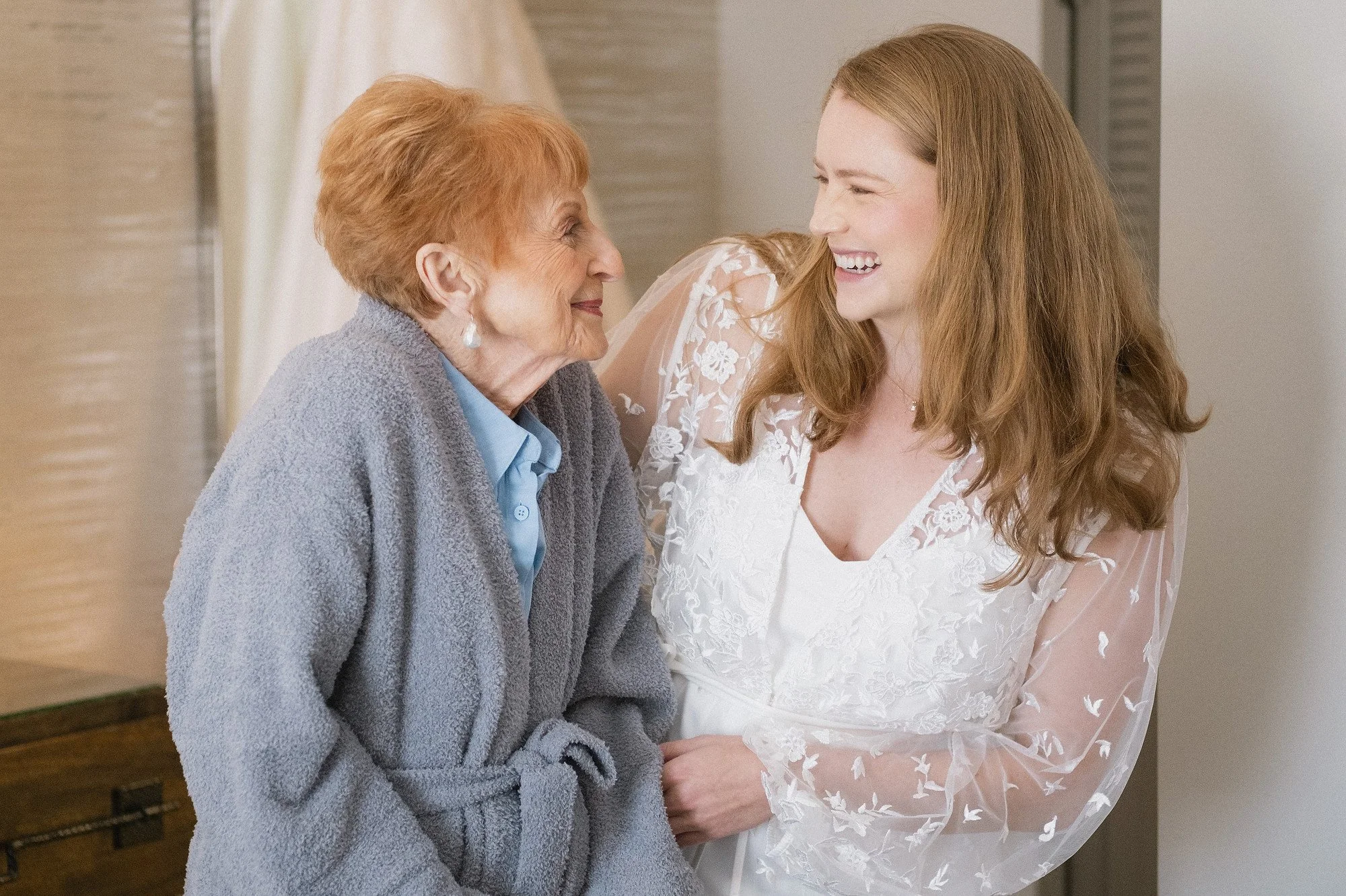 A young woman in a white dress laughing and gazing at an older woman in a grey robe who is smiling back at her.