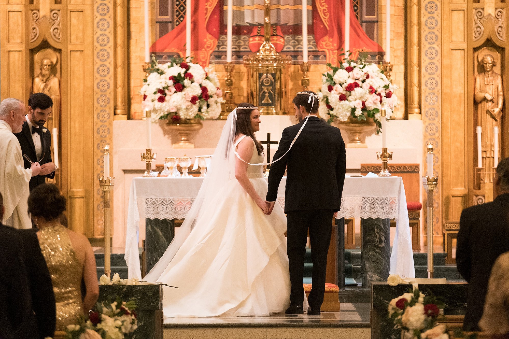 A bride and groom holding hands during a wedding ceremony inside a church with floral arrangements and religious icons in the background.