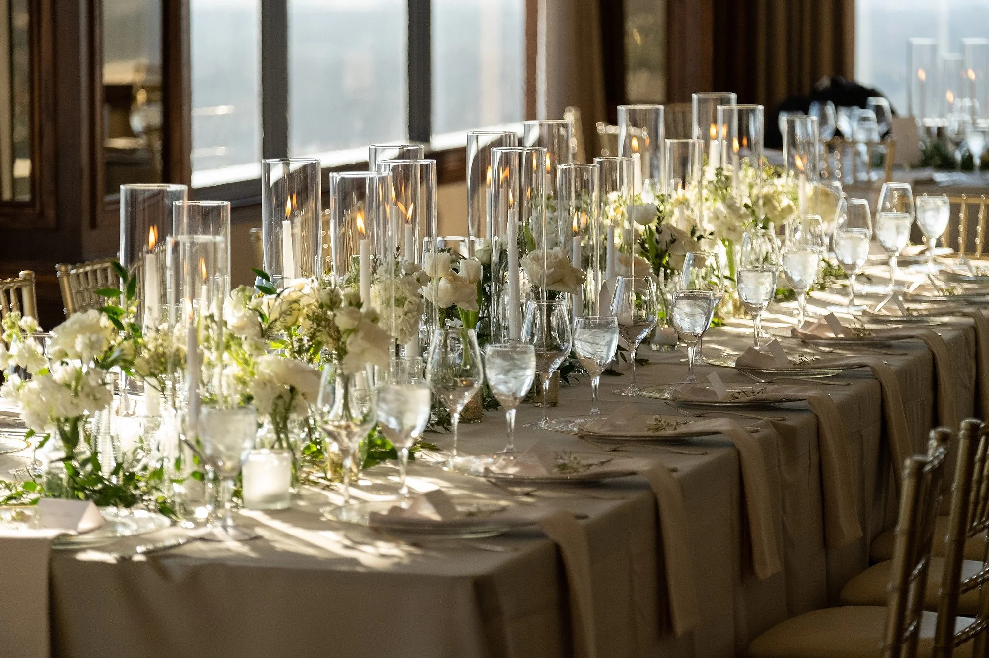 Elegant banquet table with white floral centerpieces and lit candles, set for a formal event or wedding in a sunlit room.