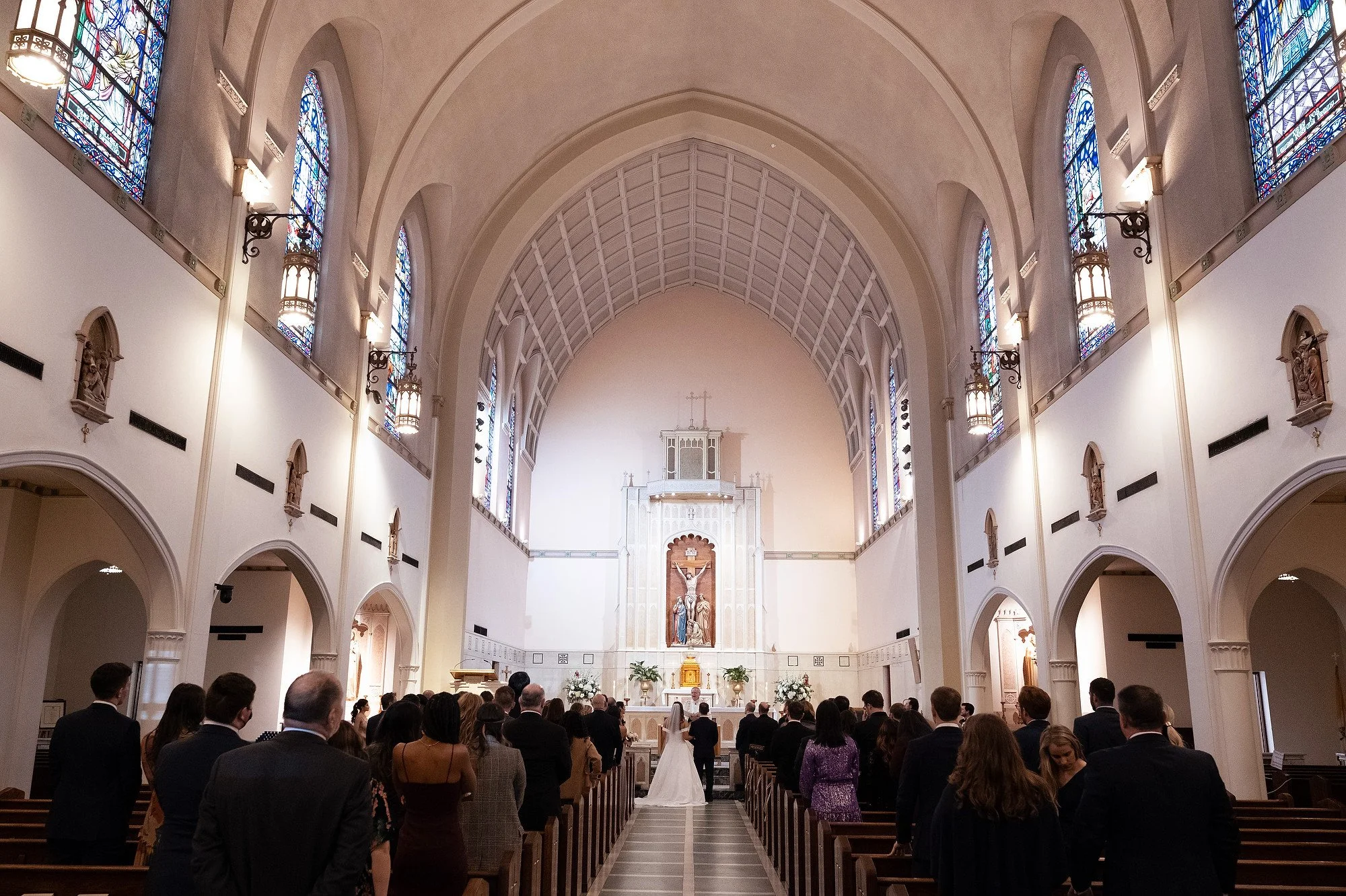 A wedding ceremony taking place inside a large church with high vaulted ceilings and stained glass windows. The bride and groom are standing at the altar facing the officiant, with guests seated in pews on either side.