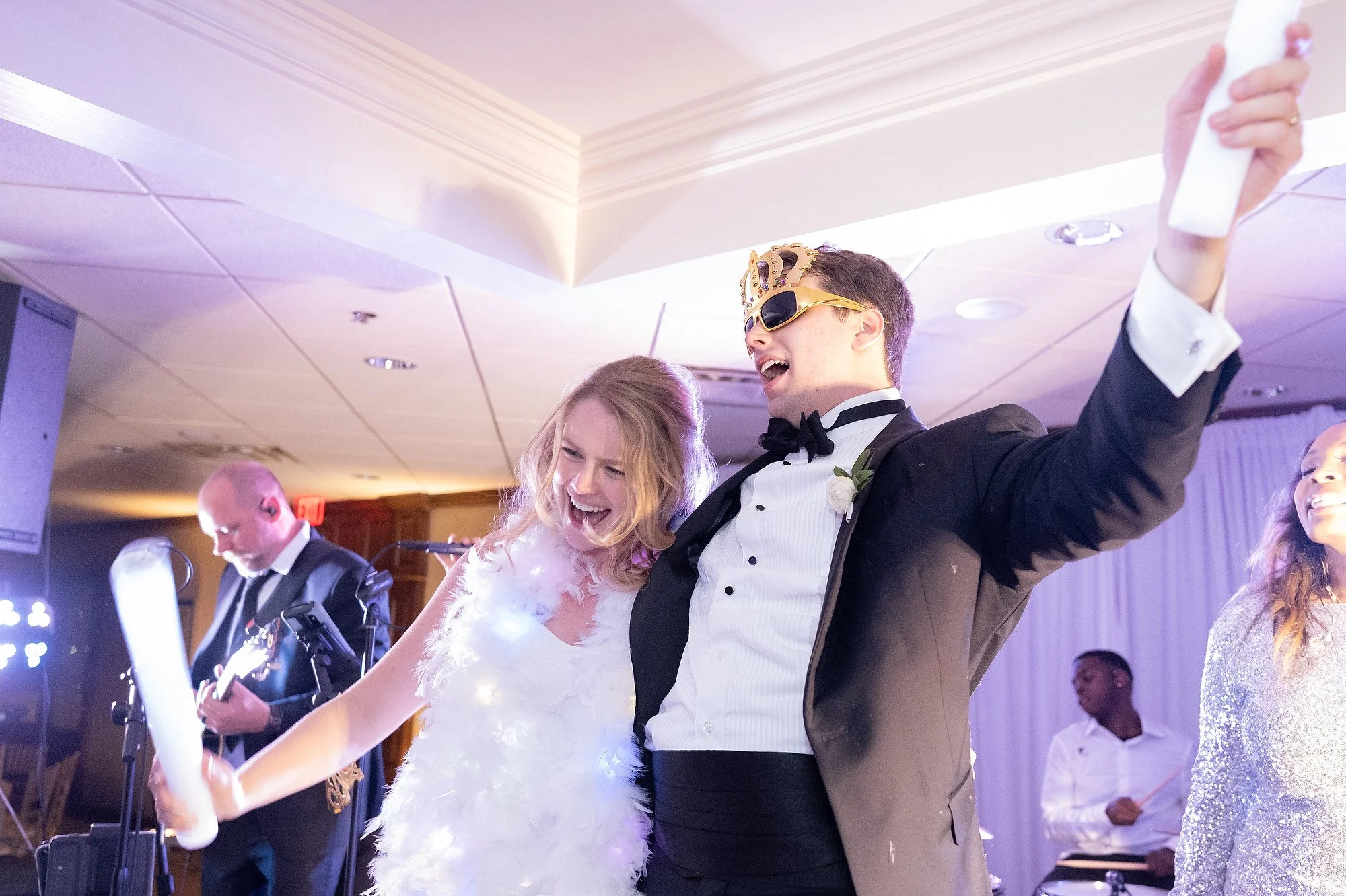 Celebrating couple at wedding reception, with woman in white feather dress and man in tuxedo wearing tiara and sunglasses, dancing with others in the background.