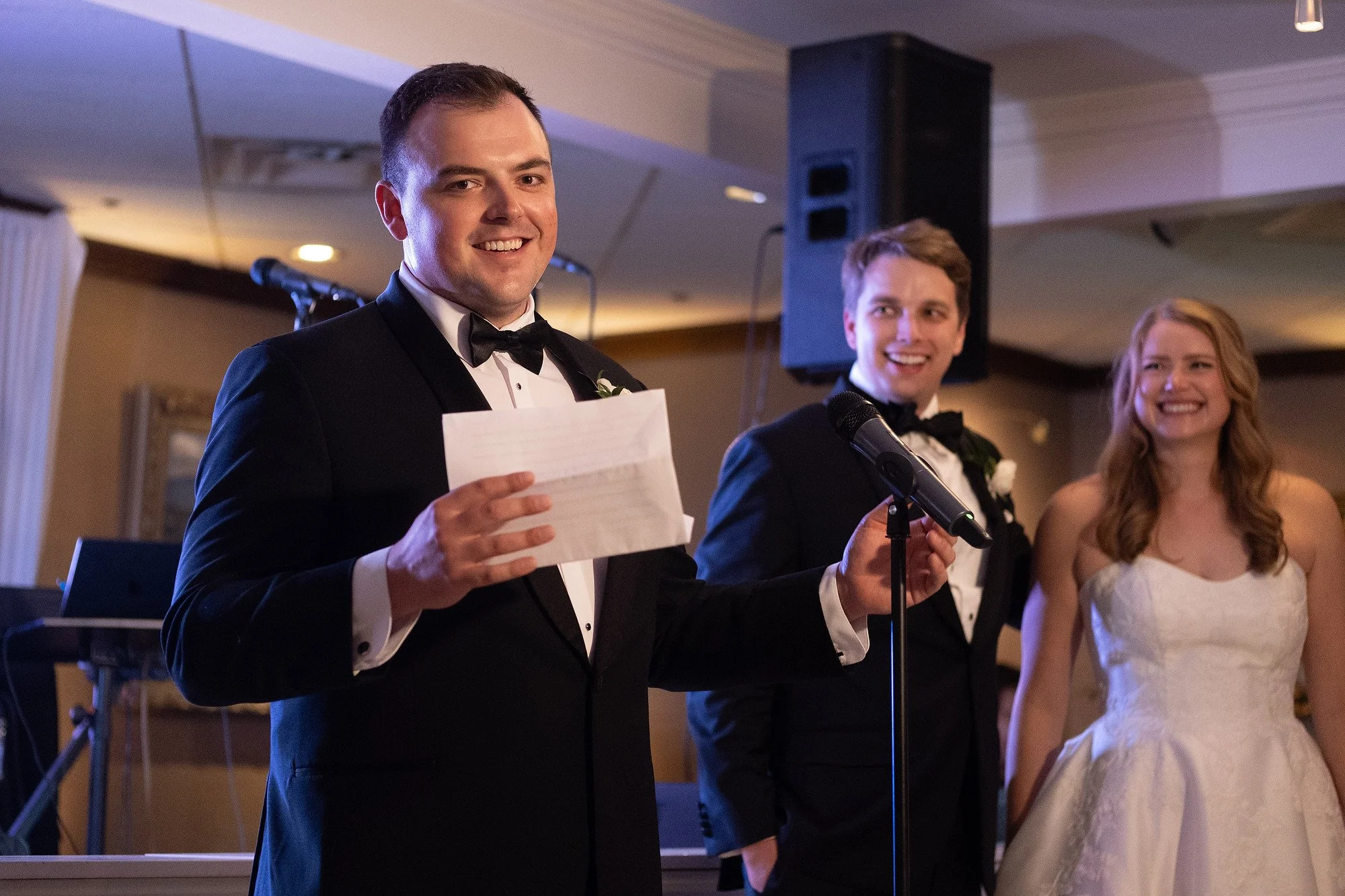 A man in a tuxedo giving a speech at a wedding reception, with two people smiling behind him, one man in a tuxedo and one woman in a wedding dress.