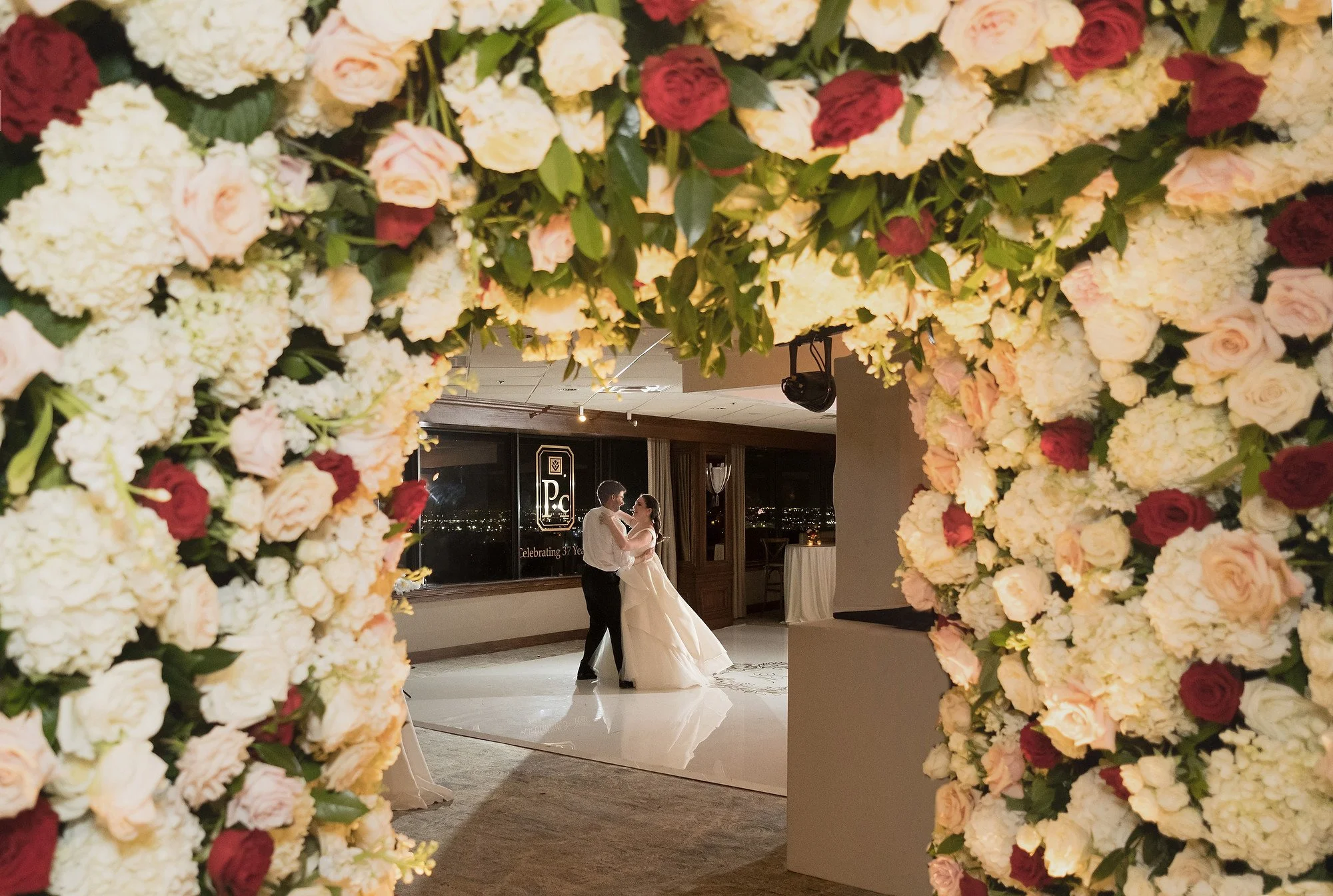 A bride and groom dancing at their wedding reception, viewed through a floral arch of white, cream, blush, and burgundy flowers.