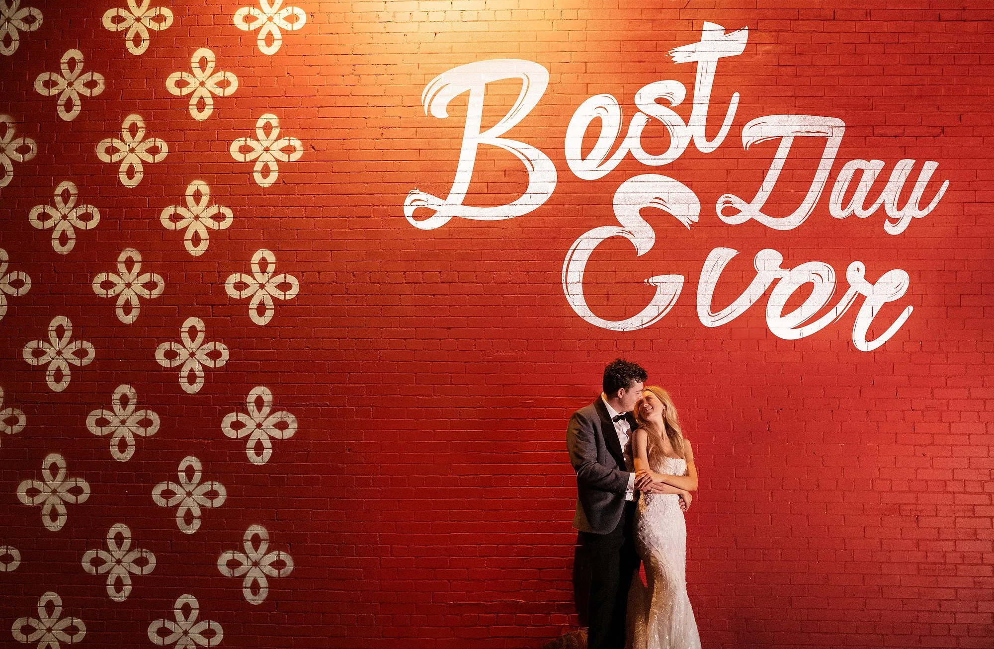 Couple in wedding attire standing in front of a red brick wall with large white text saying 'Best Day Ever' and decorative white floral patterns.