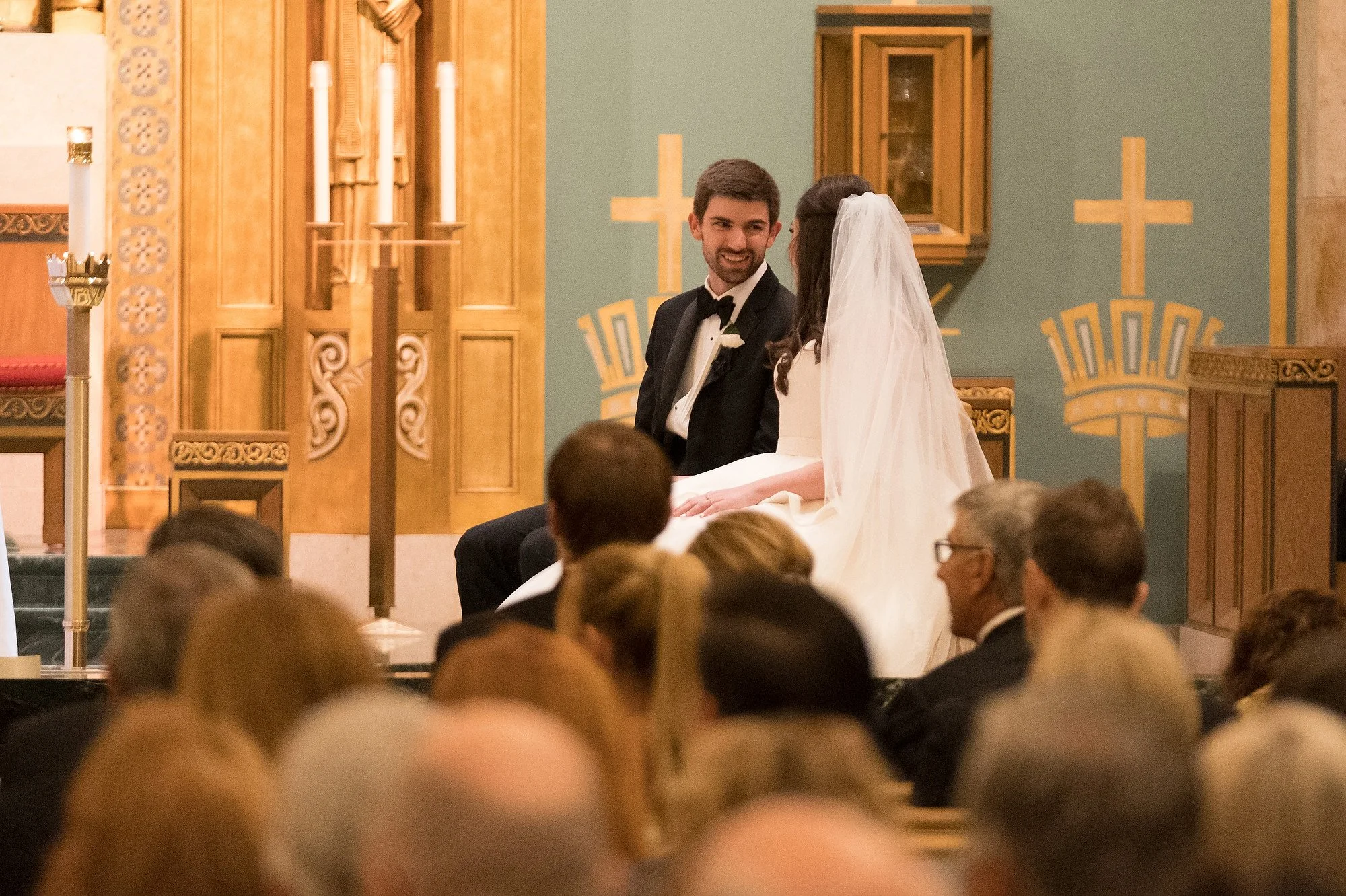 A bride and groom sitting on a church altar during a wedding ceremony, with guests seated in front of them.