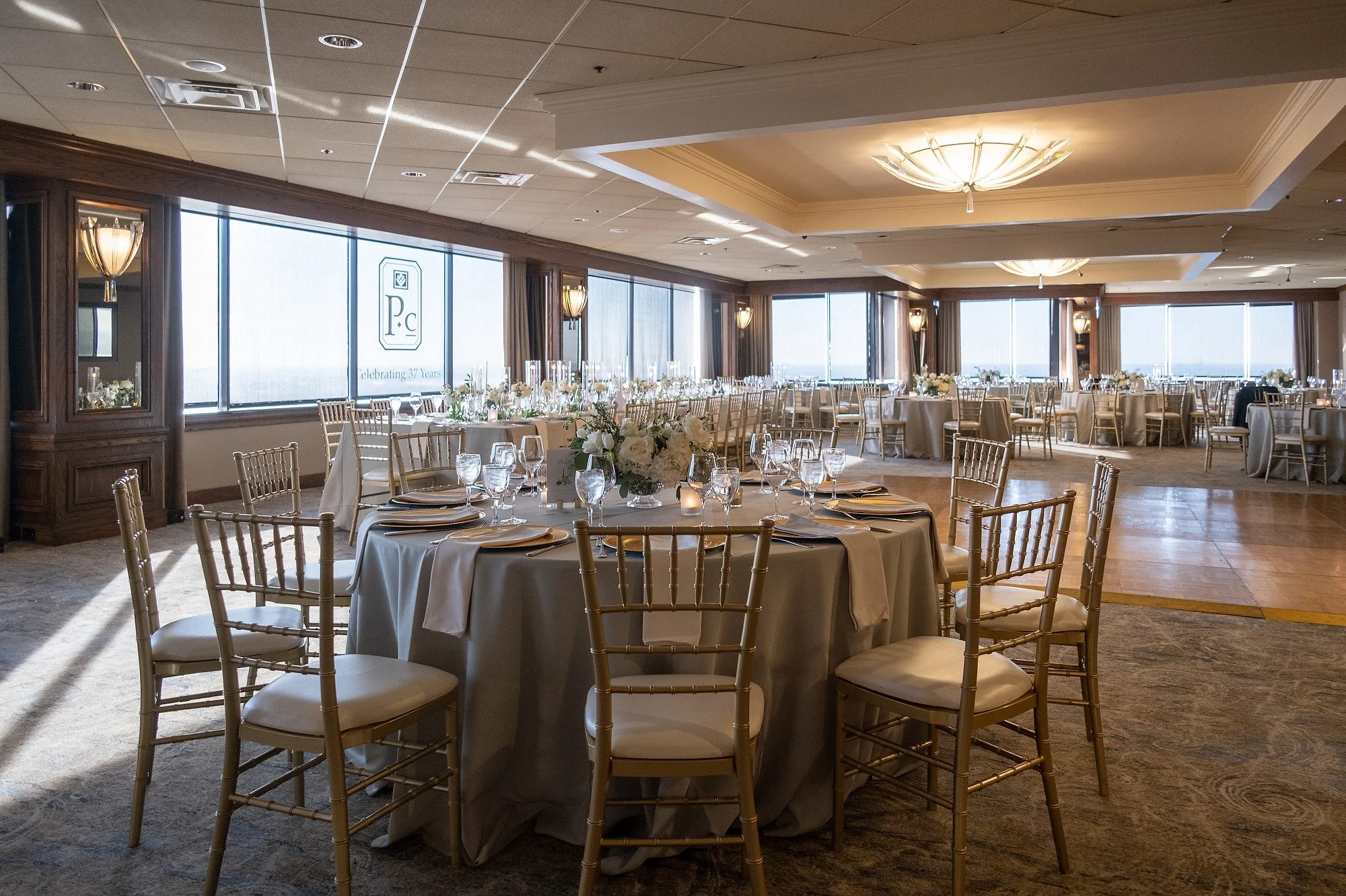 Elegant banquet hall decorated for a celebration with round tables covered in beige tablecloths, floral centerpieces, candle decorations, and surrounded by white and gold chairs. Large windows let in natural light, and the ceiling has modern light fi