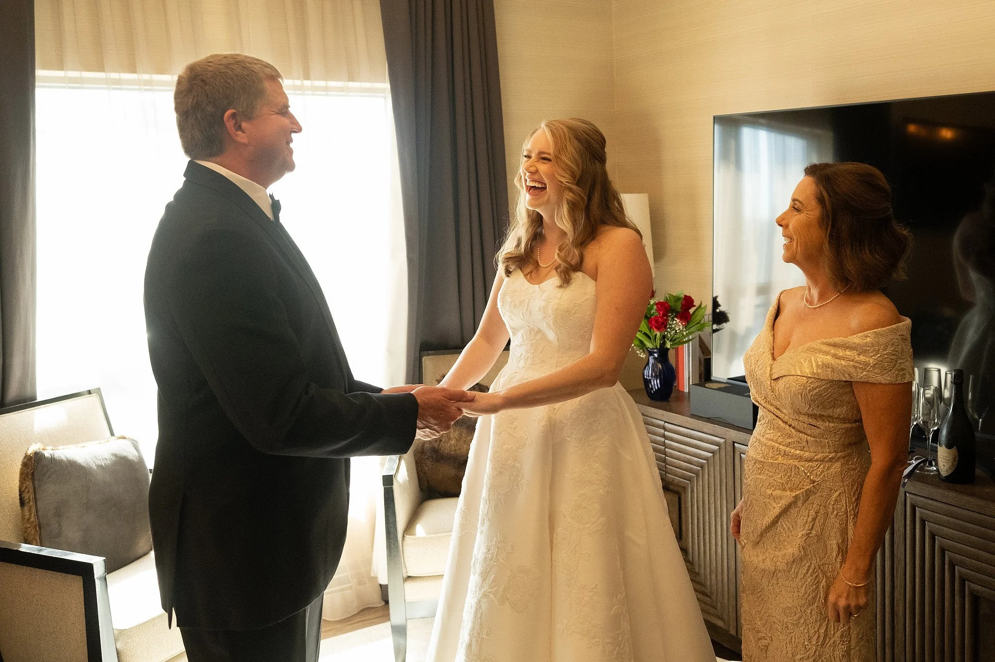 A groom and bride holding hands, smiling at each other during a wedding ceremony, with a woman in a gold dress standing nearby in a well-lit room.