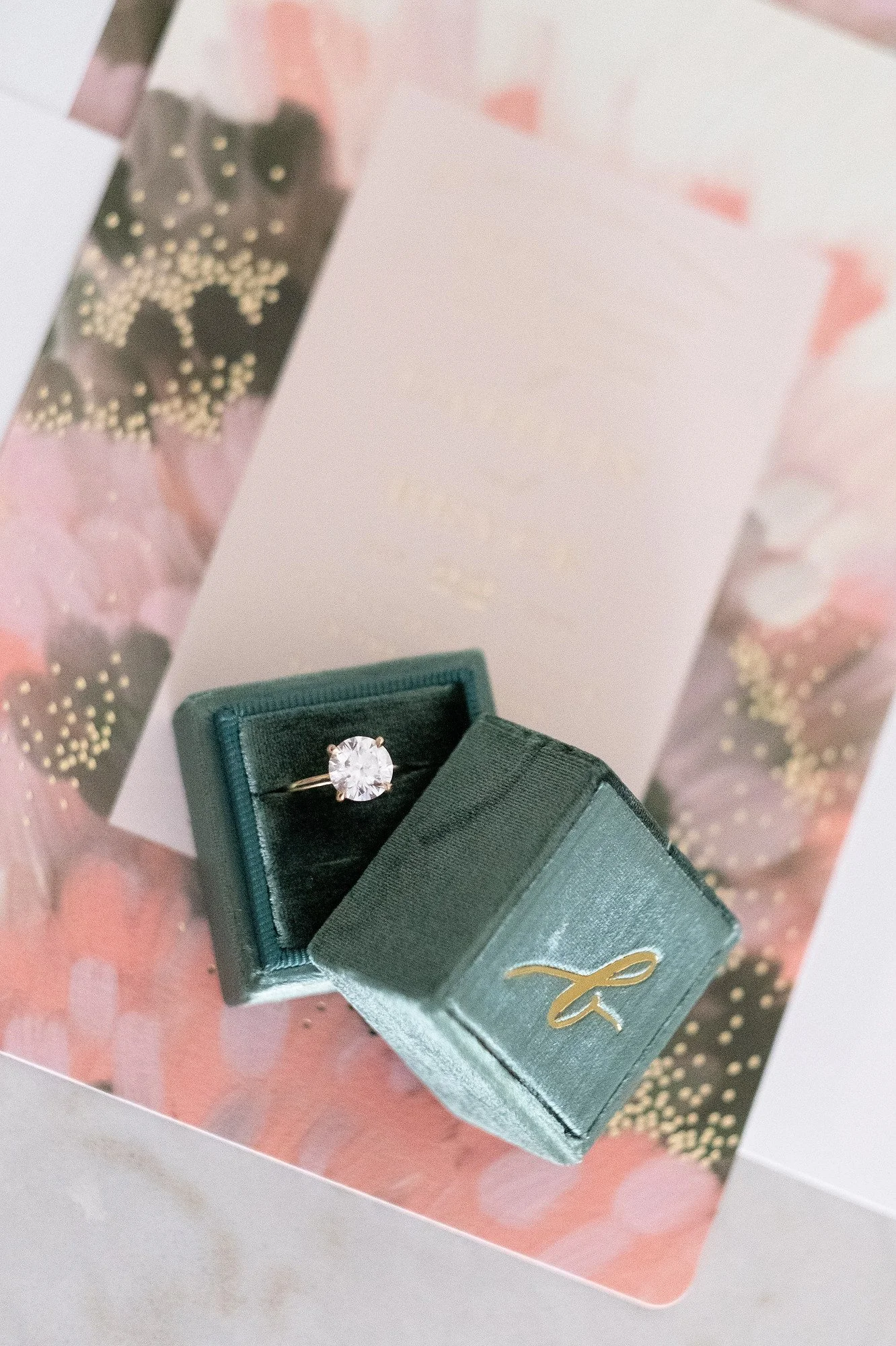 A diamond engagement ring in a green velvet ring box, placed on a decorative card or paper with pink and black floral print and gold accents.