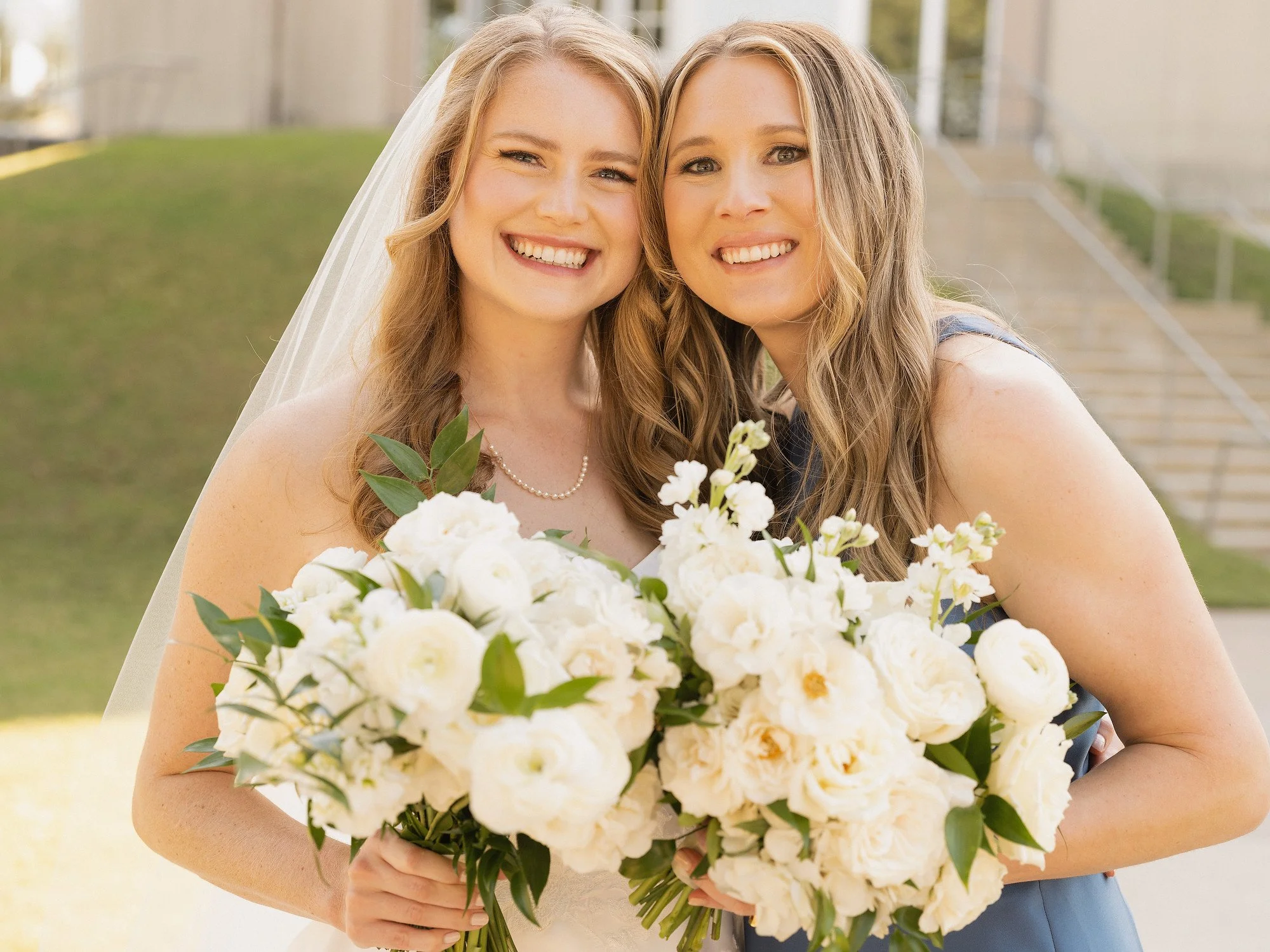 Two smiling women, one in a wedding dress and veil, holding large bouquets of white flowers, standing outdoors on steps.
