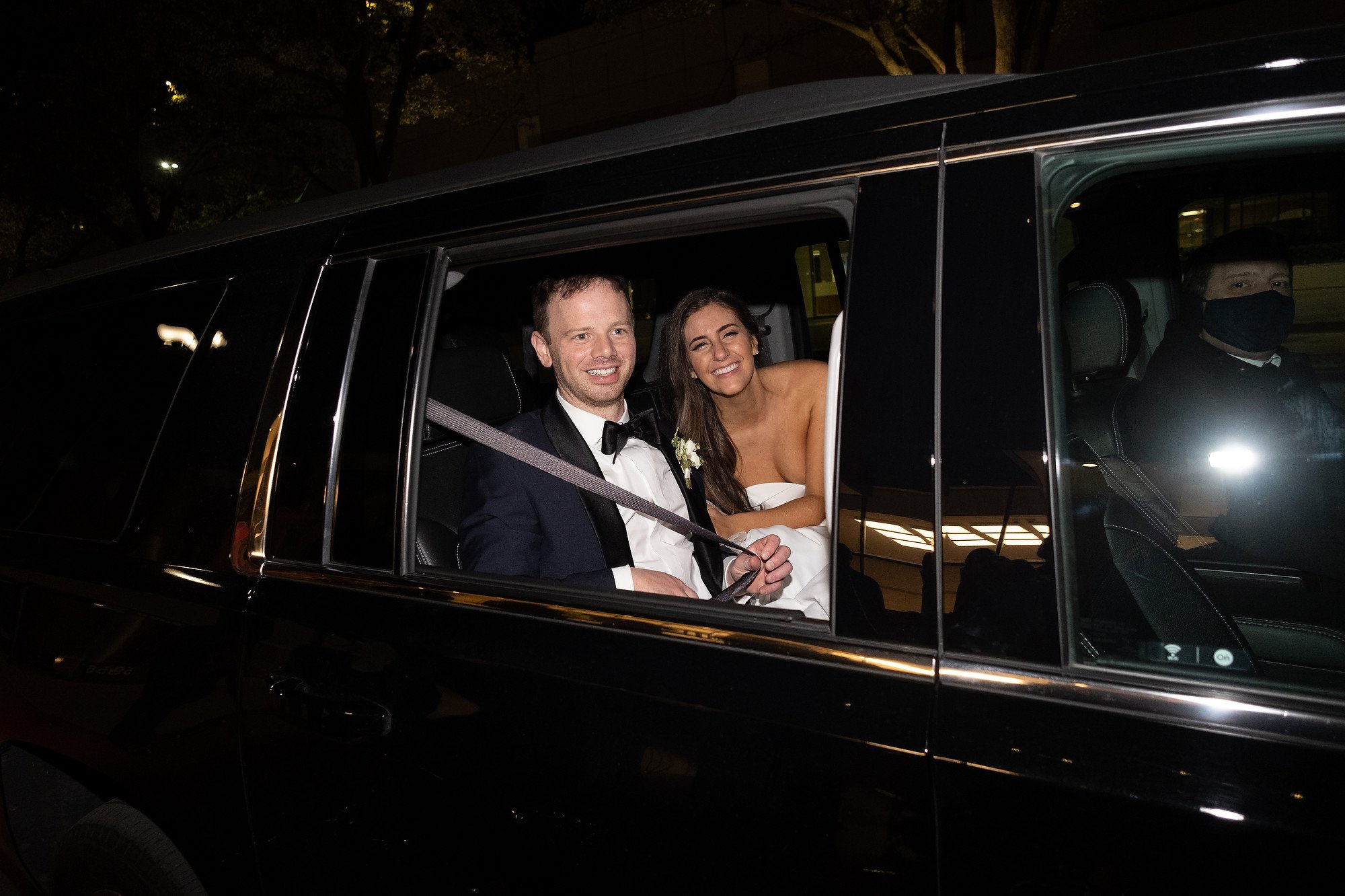 Bride and groom sitting in the back of a black limousine during their wedding, with the groom wearing a tuxedo and the bride in a strapless white dress, both smiling.