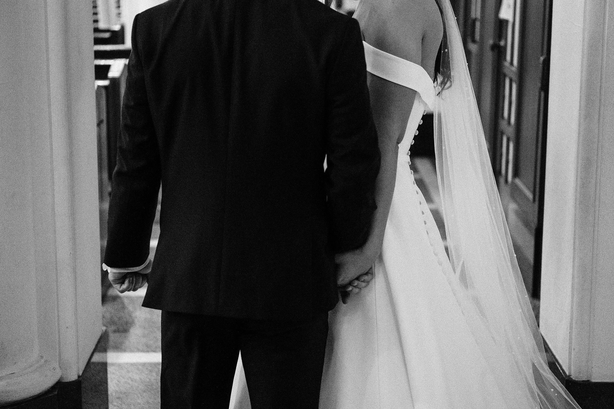 A black and white photo of a bride and groom holding hands, with the groom in a suit and the bride in a wedding dress with a veil.