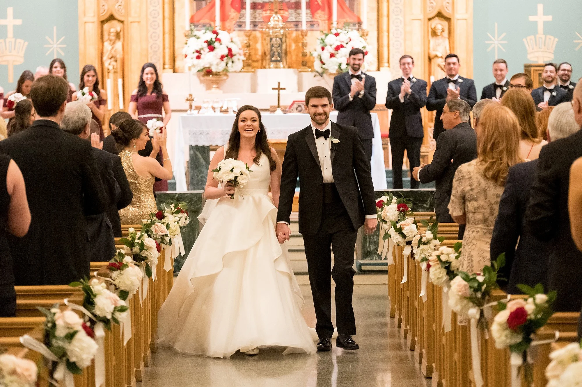 A newlywed couple walking down the aisle hand in hand inside a decorated church, smiling with guests clapping and celebrating around them.
