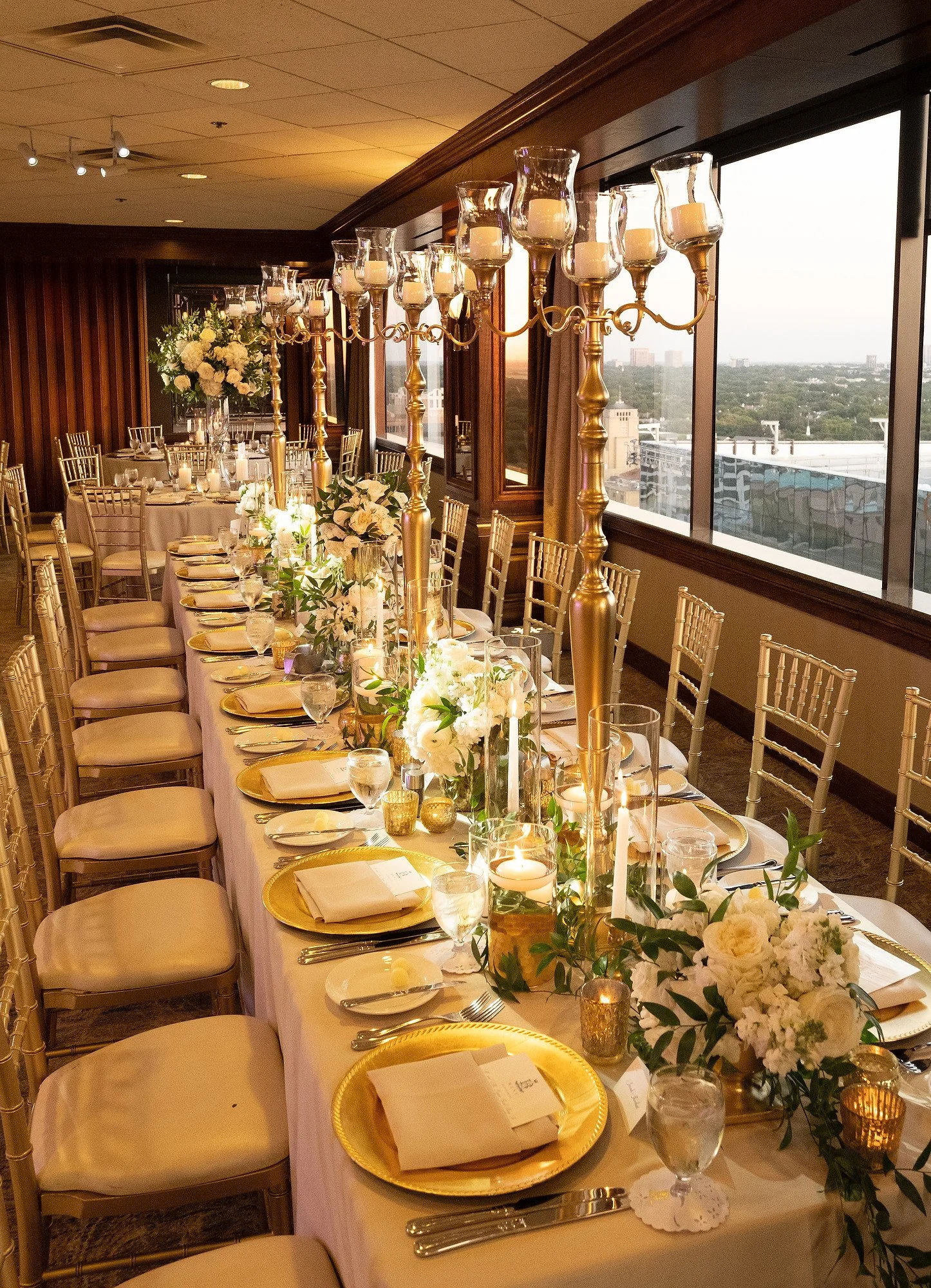 Elegant indoor wedding reception table decorated with gold chargers, white napkins, glassware, floral arrangements, candles, and tall gold candle holders. Large windows reveal a city view at sunset.