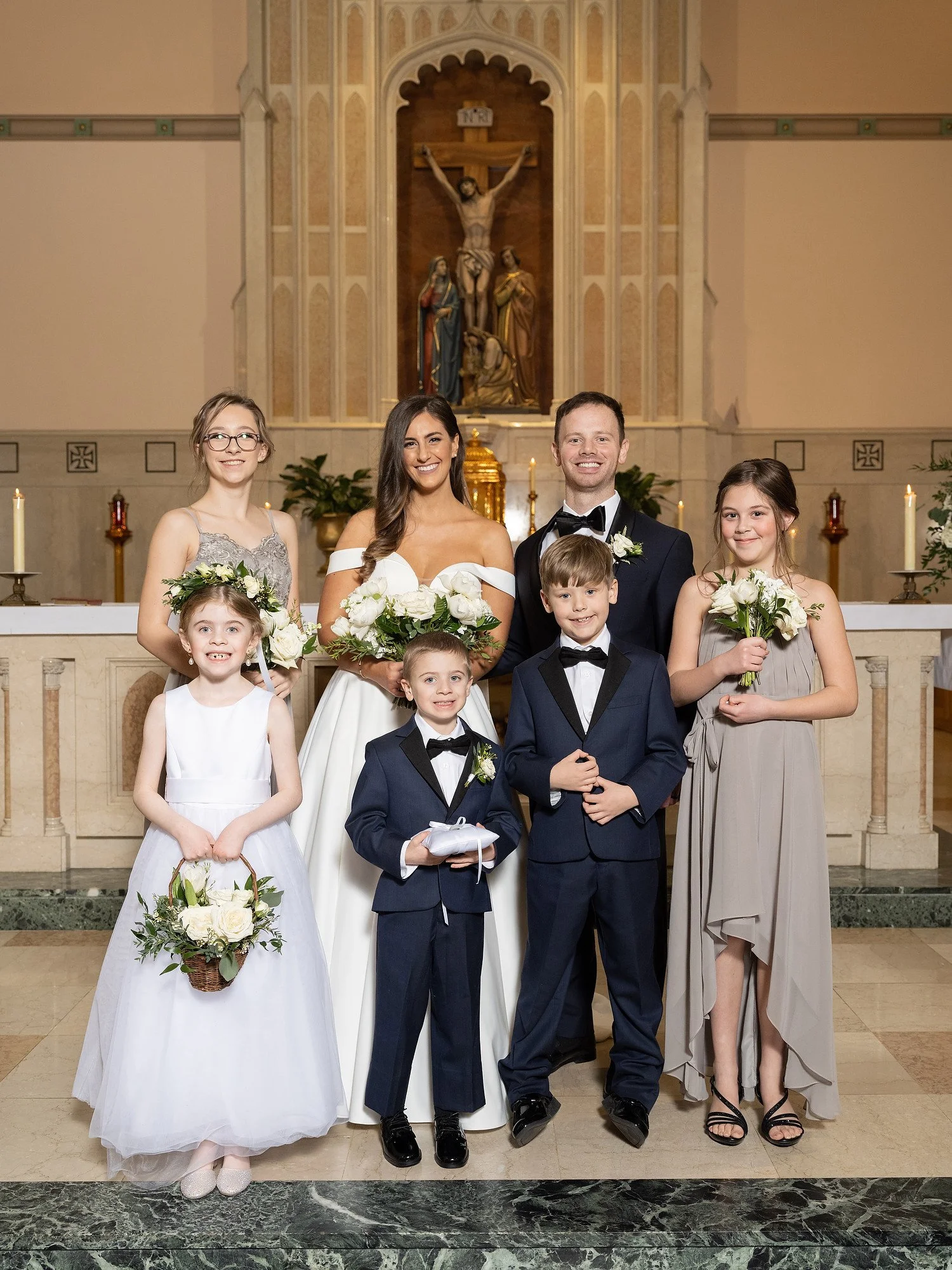 A wedding party in a church with a crucifix and religious statues in the background. The group includes a bride, groom, and five children, all in formal attire, smiling and holding flowers or gifts.