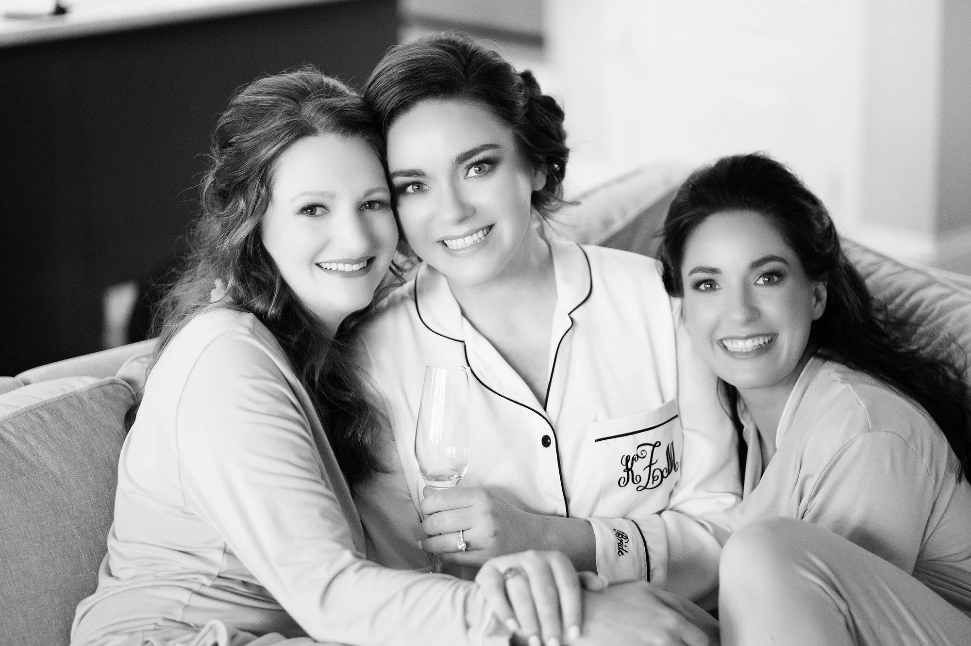 Three women in pajamas smiling happily, sitting close together on a couch, with one woman holding a glass of champagne.