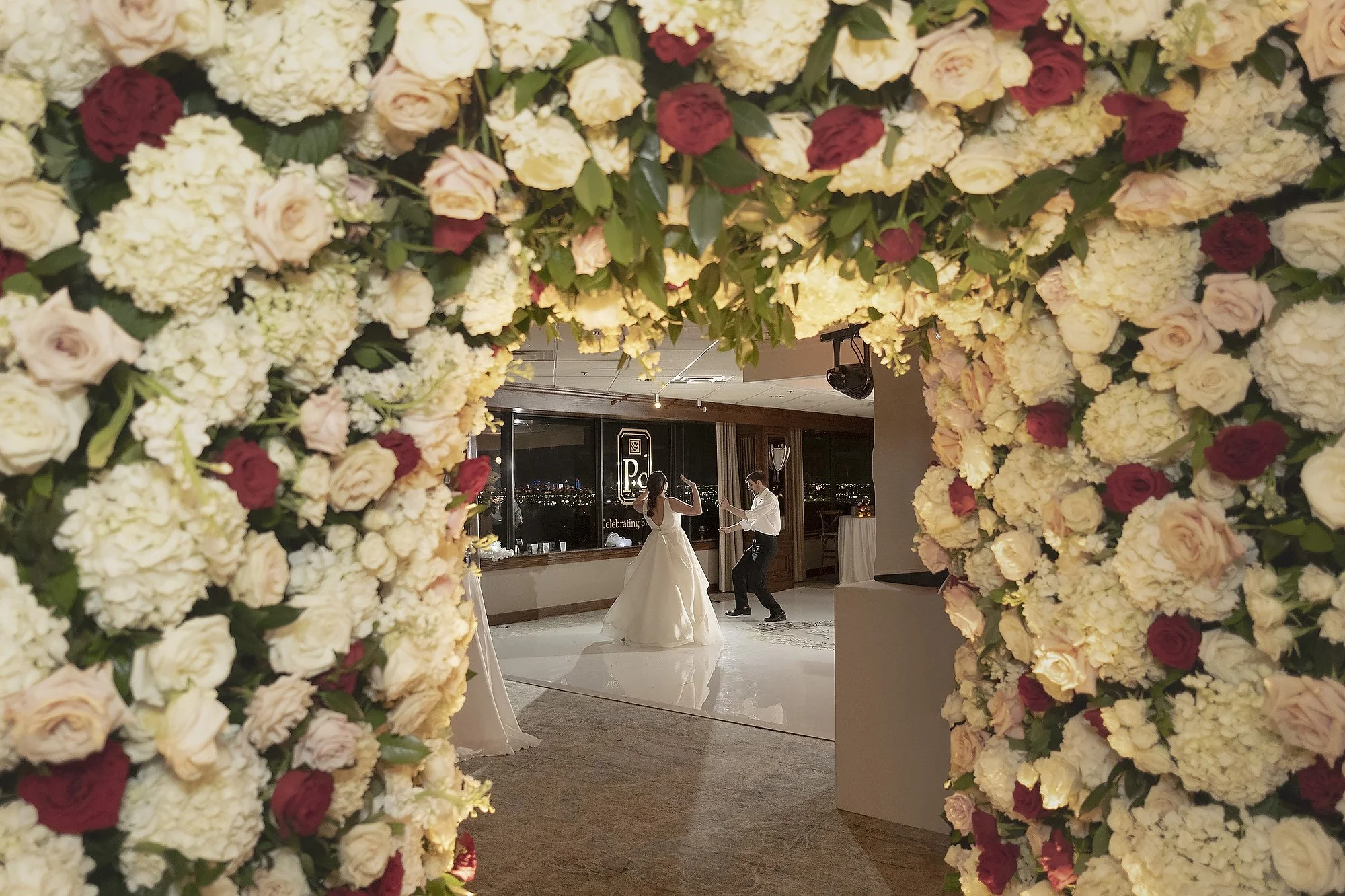 A bride and groom dancing in a decorated wedding venue, viewed through a large archway of cream, blush, and red roses and hydrangeas.