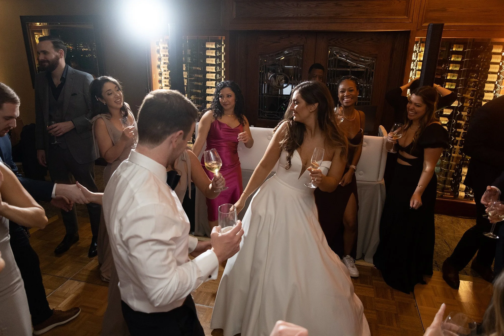 People dancing and celebrating at a wedding reception, with women in formal dresses and men in suits, holding glasses of wine or champagne.
