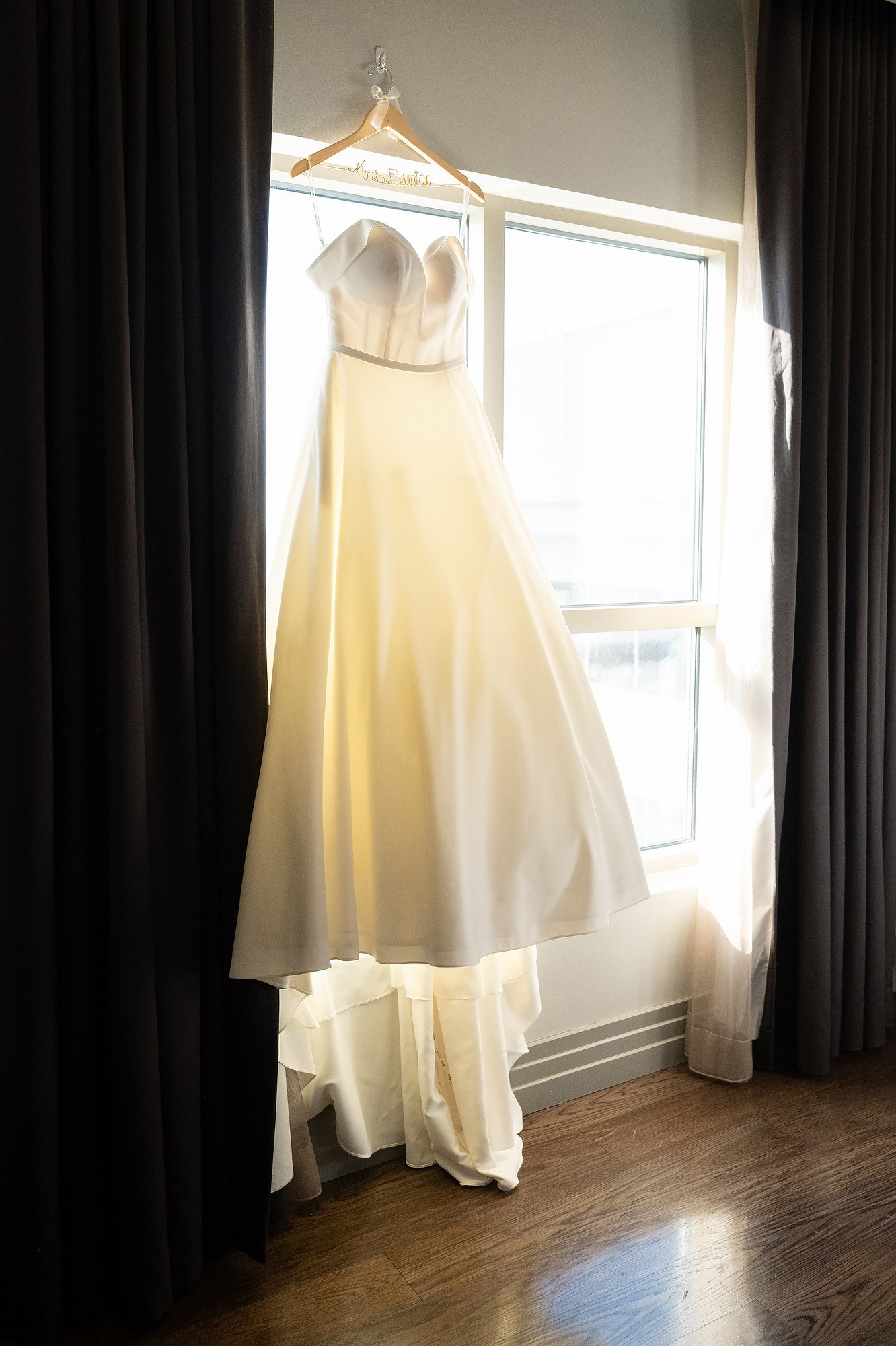 A white wedding dress hanging on a hanger in front of a window, with black and white curtains on each side.