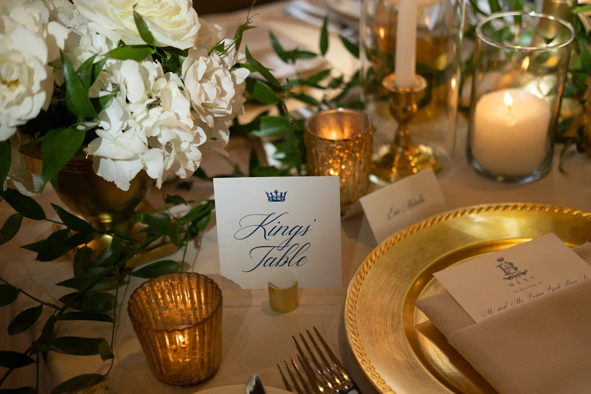 A formal dinner table decorated with white flowers, green leaves, lit candles, and gold accents. A sign reads 'Kings' Table', indicating a special seating arrangement.