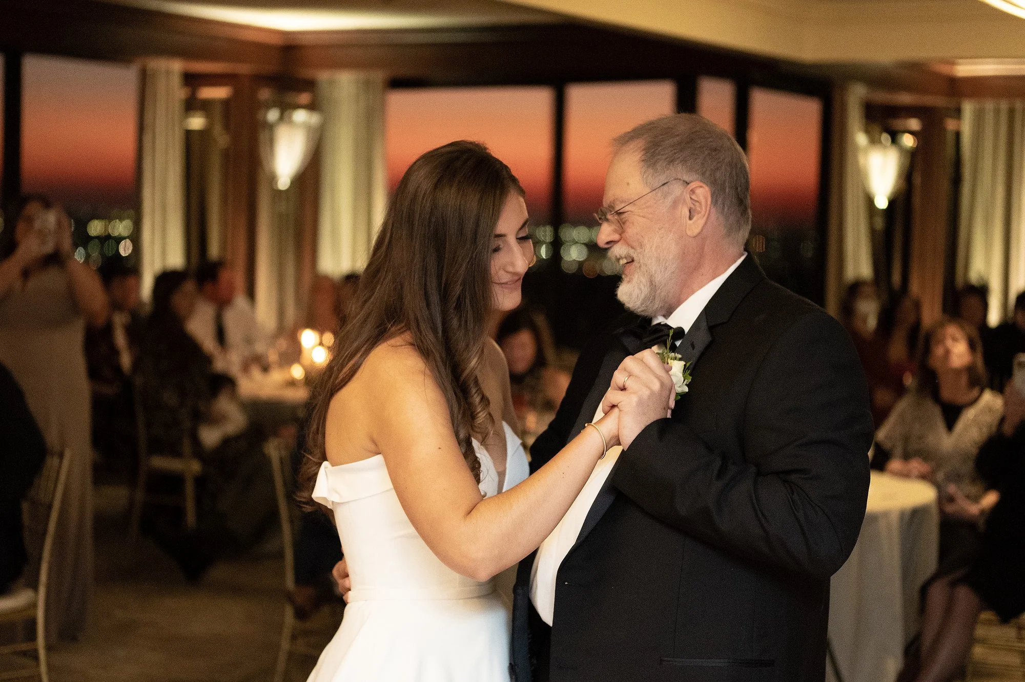 A bride and her father share a dance at her wedding reception in a decorated banquet room during sunset.