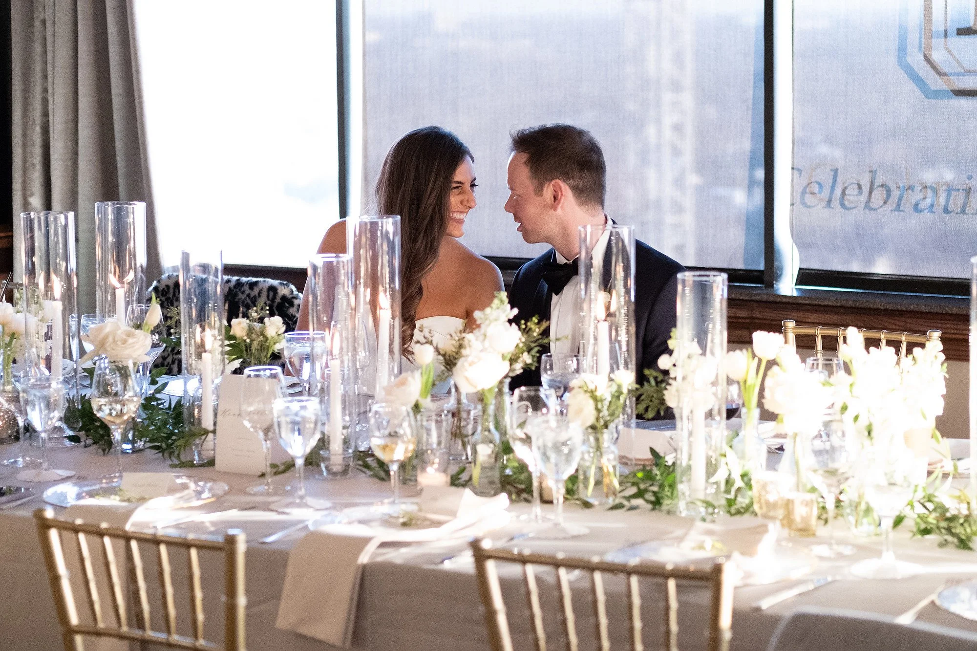 A bride and groom sitting at a decorated wedding reception table, smiling at each other.