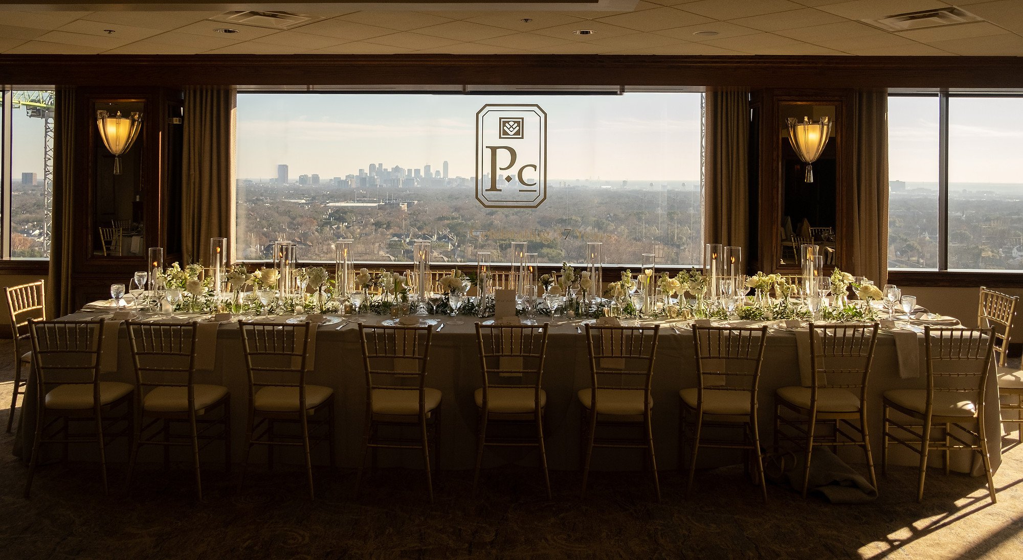 Elegant banquet table with white tablecloth, floral centerpieces, and tall candles, set in front of large windows overlooking a city skyline during daytime. The windows have a logo with the initials P.C.