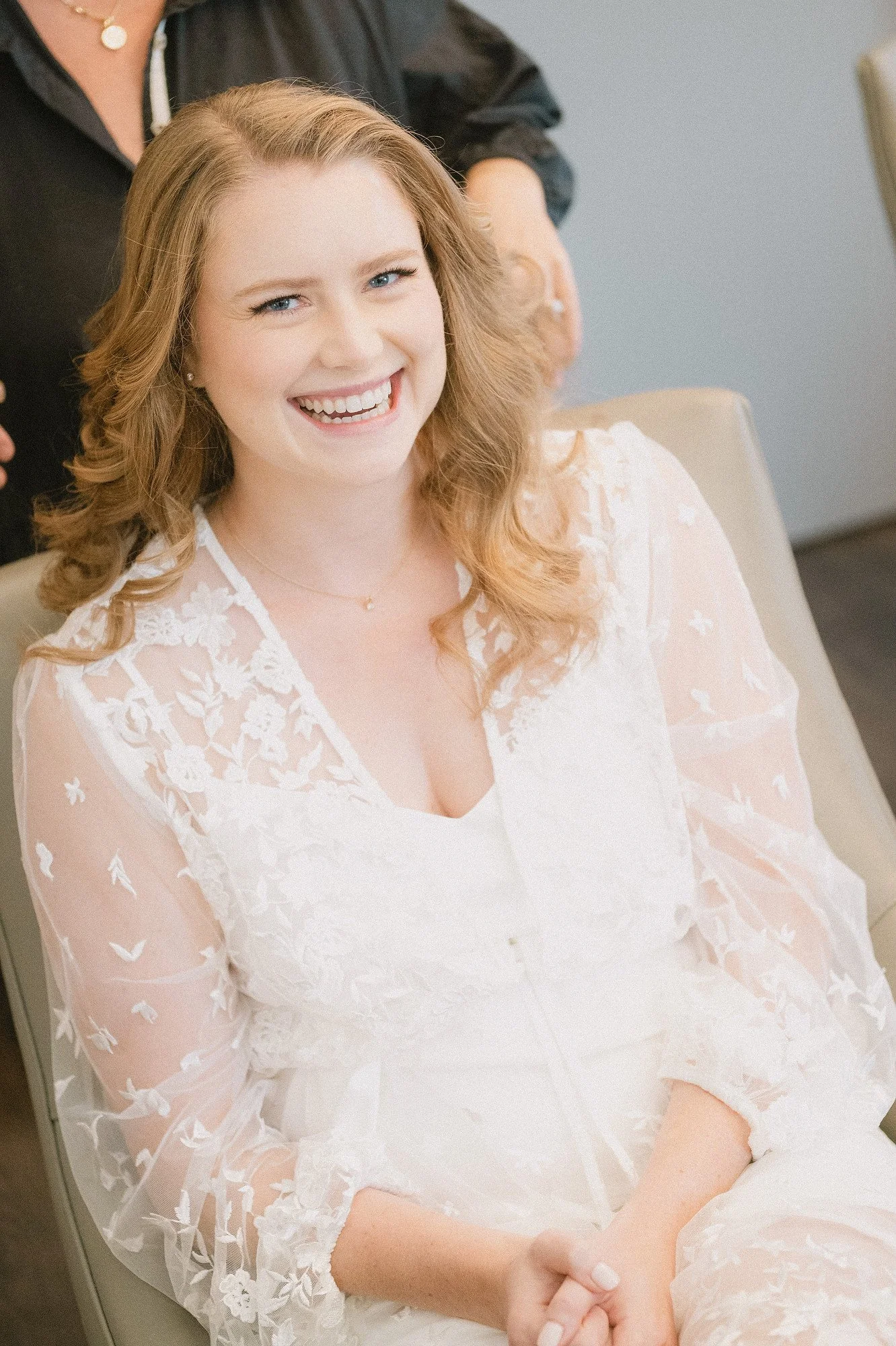 A woman with light skin, red hair, and blue eyes, smiling, wearing a white, lace dress, sitting in a chair.