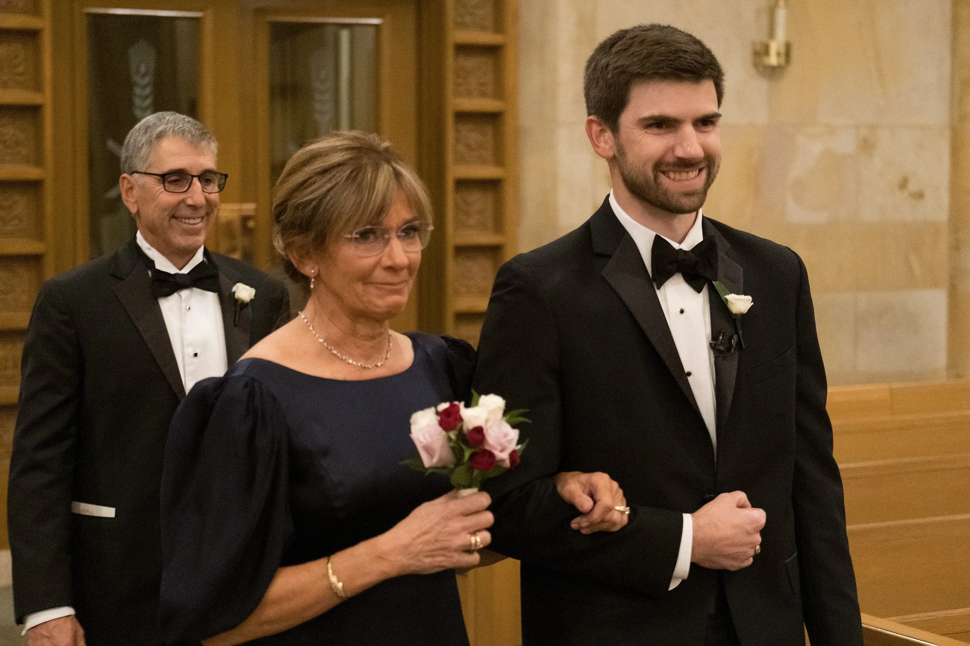 A wedding ceremony featuring three people dressed in formal tuxedos and dresses, with the officiant leading the bride and groom down the aisle in a church setting.