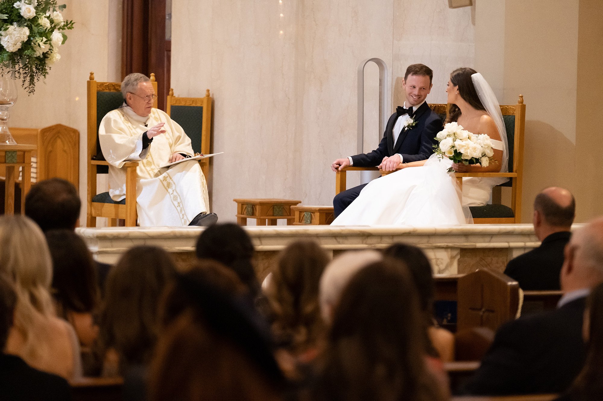 A bride and groom sitting on thrones during their wedding ceremony, facing a priest who is officiating the service, in a church setting with guests in attendance.