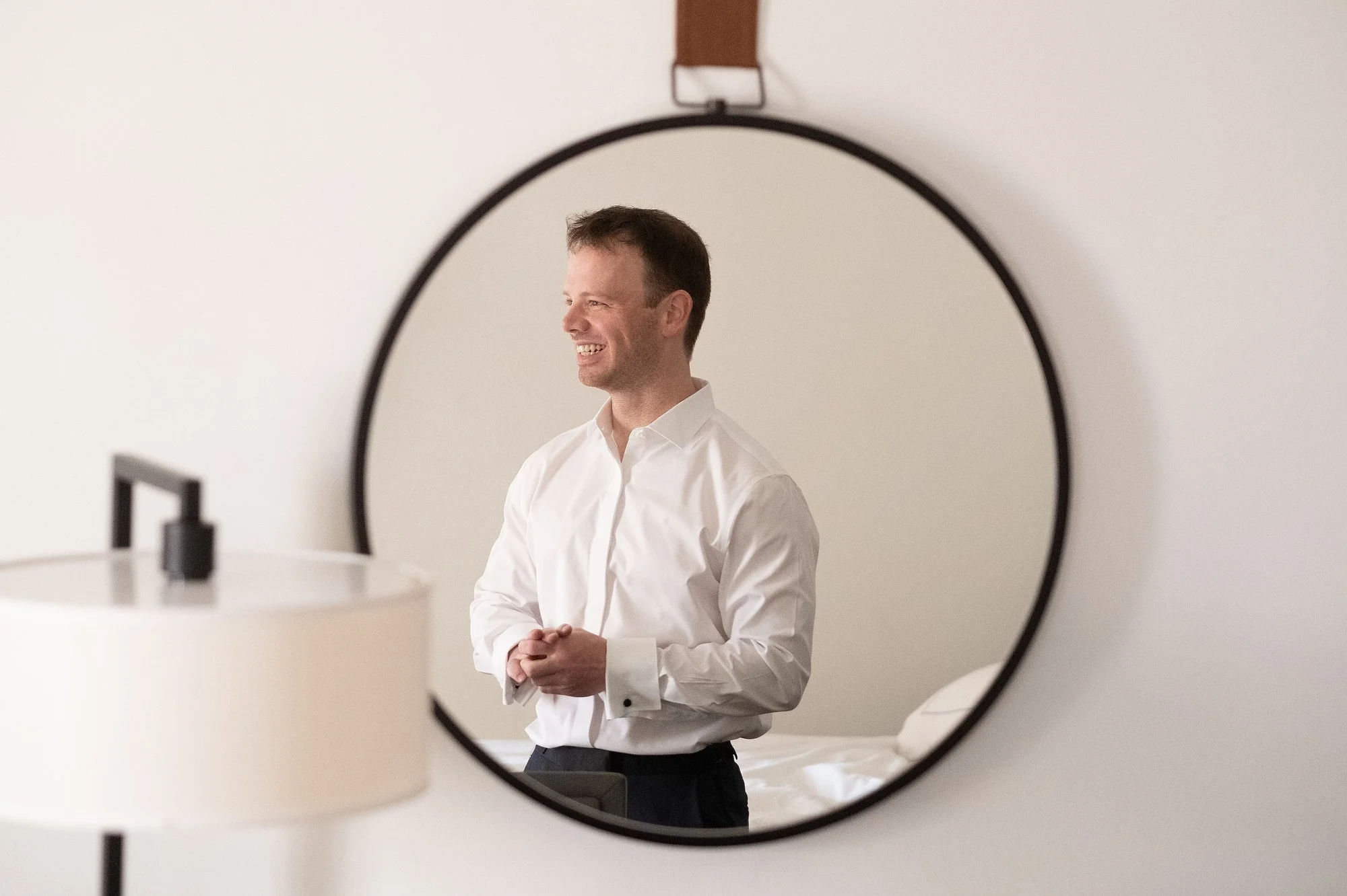 A man in a white shirt smiling, reflected in a circular mirror in a minimalistic room with neutral tones.