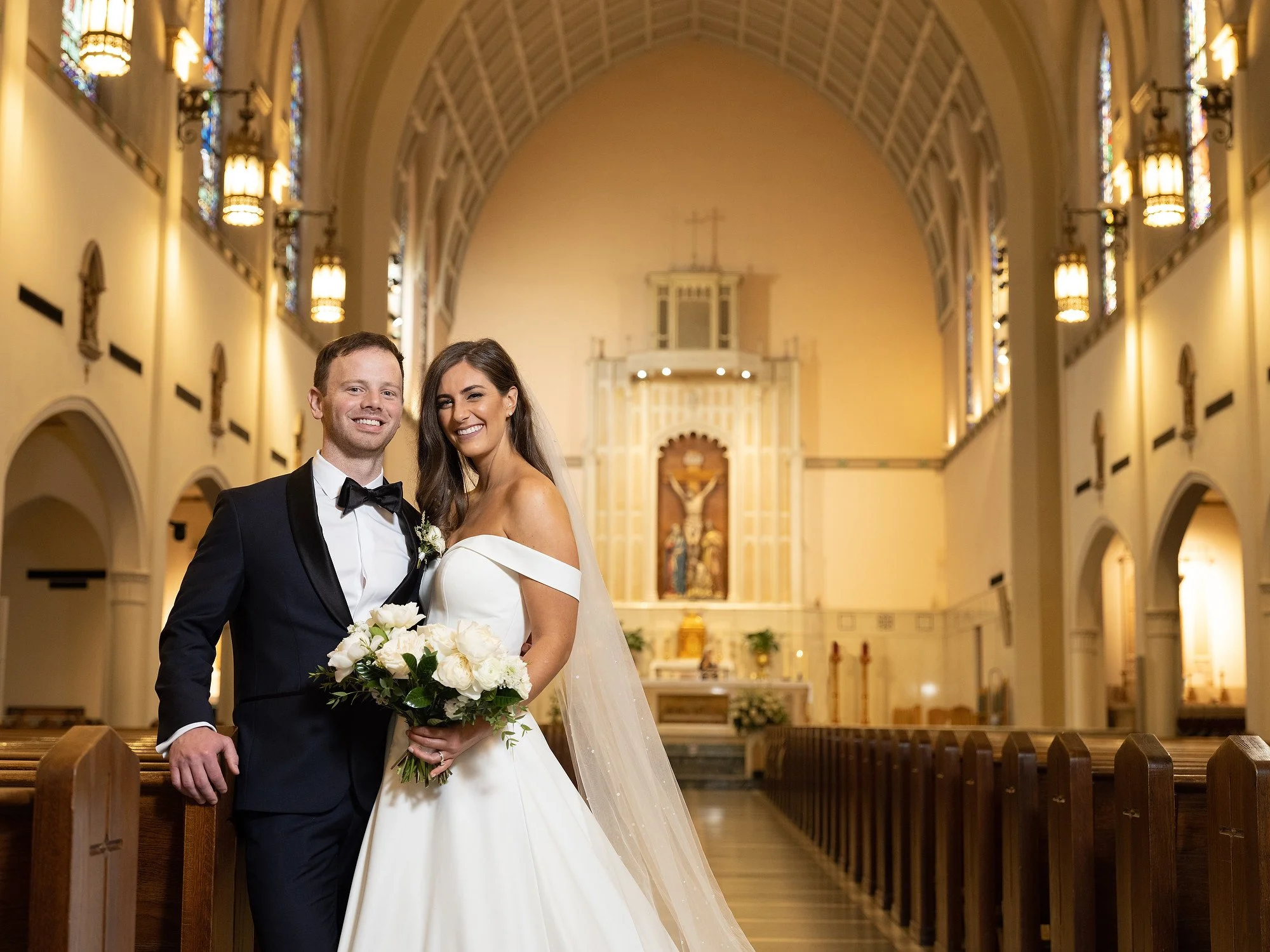 A newlywed couple dressed in wedding attire standing inside a church, smiling. The groom is in a black tuxedo with a bow tie, and the bride is in a white gown holding a bouquet of white flowers. The church has high ceilings, stained glass windows, an