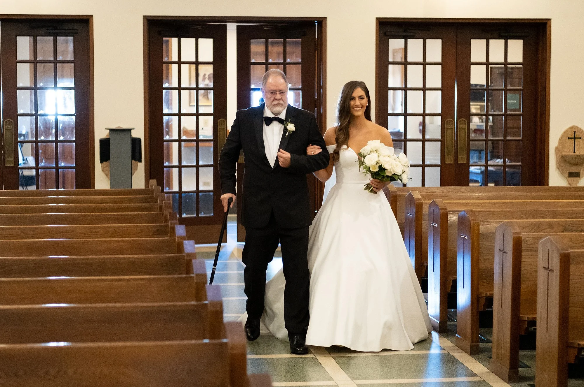 A bride in a white wedding gown holding a bouquet, walking down the aisle with an older man in a tuxedo using a cane in a church.