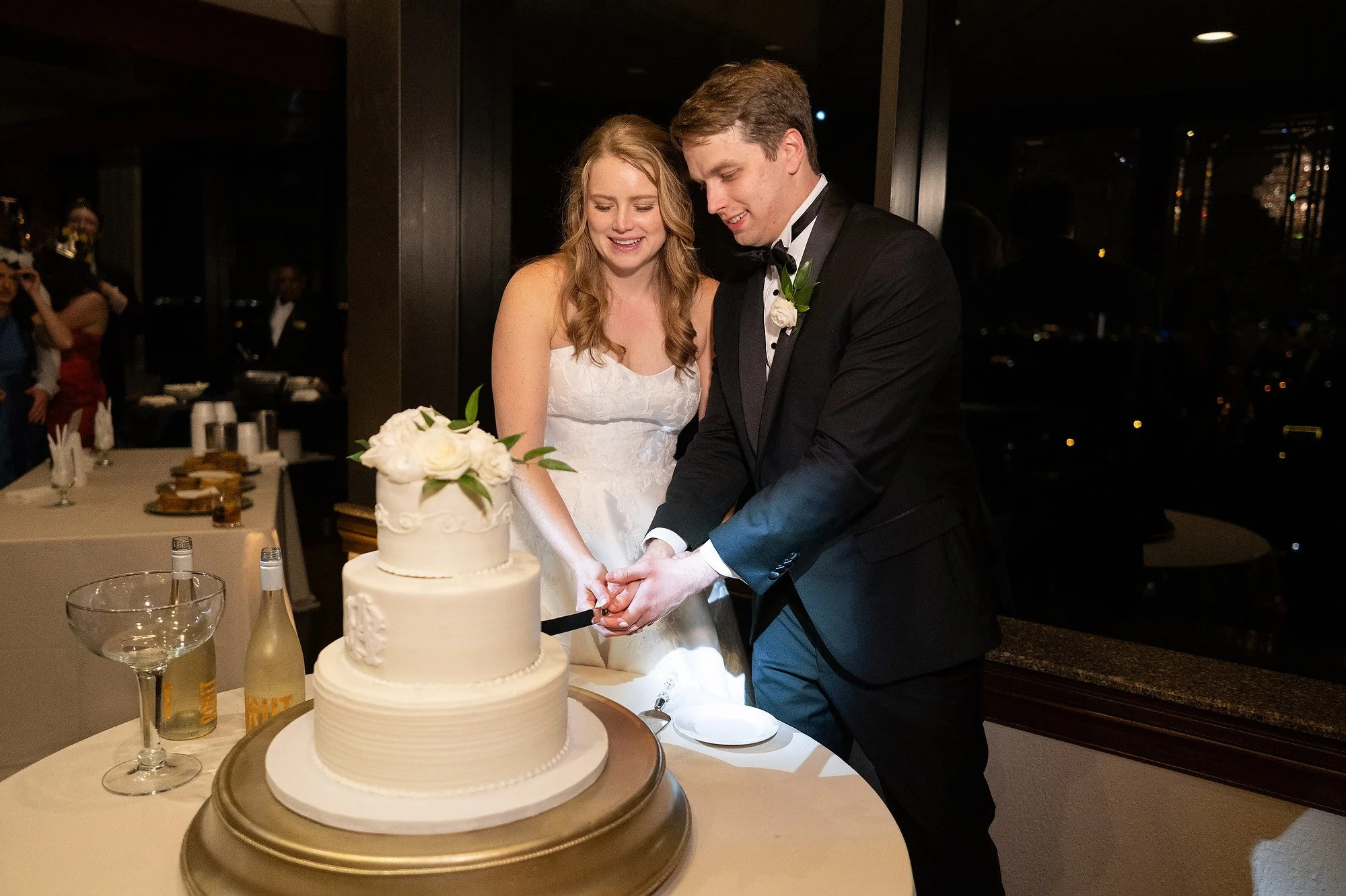 A bride and groom cutting a wedding cake together at their reception, with guests in the background.